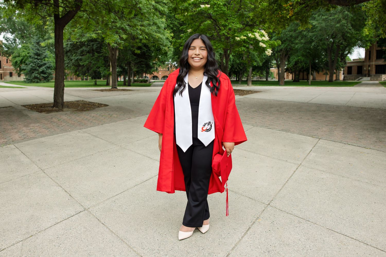 Young woman in red graduation gown and cap smiles outdoors on campus walkway surrounded by trees.