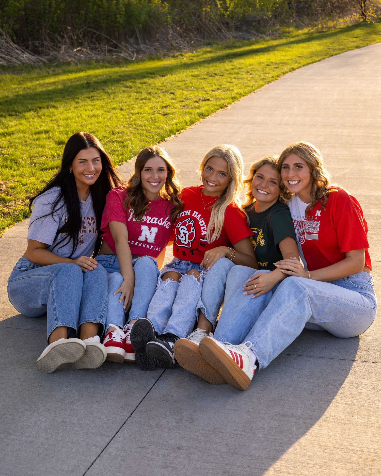 Friends in casual red tops and jeans sitting together on concrete path outdoors.