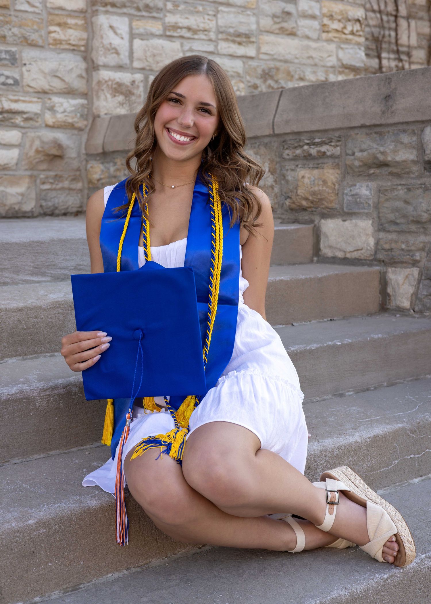 Graduate in white dress and blue stole poses playfully on stone steps for graduation photos.