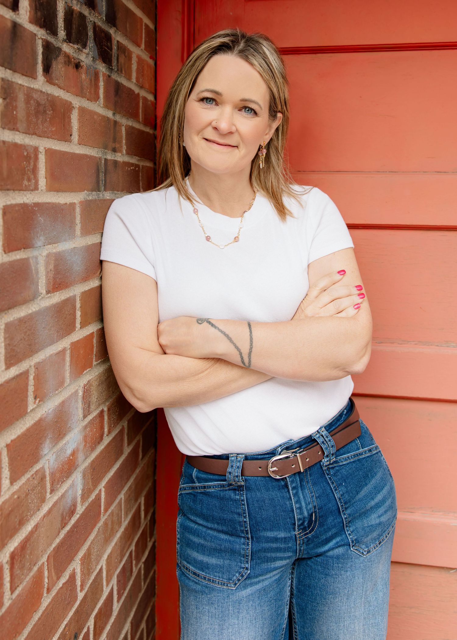 Woman in white t-shirt and jeans leaning against a red brick wall with arms crossed, smiling confidently.