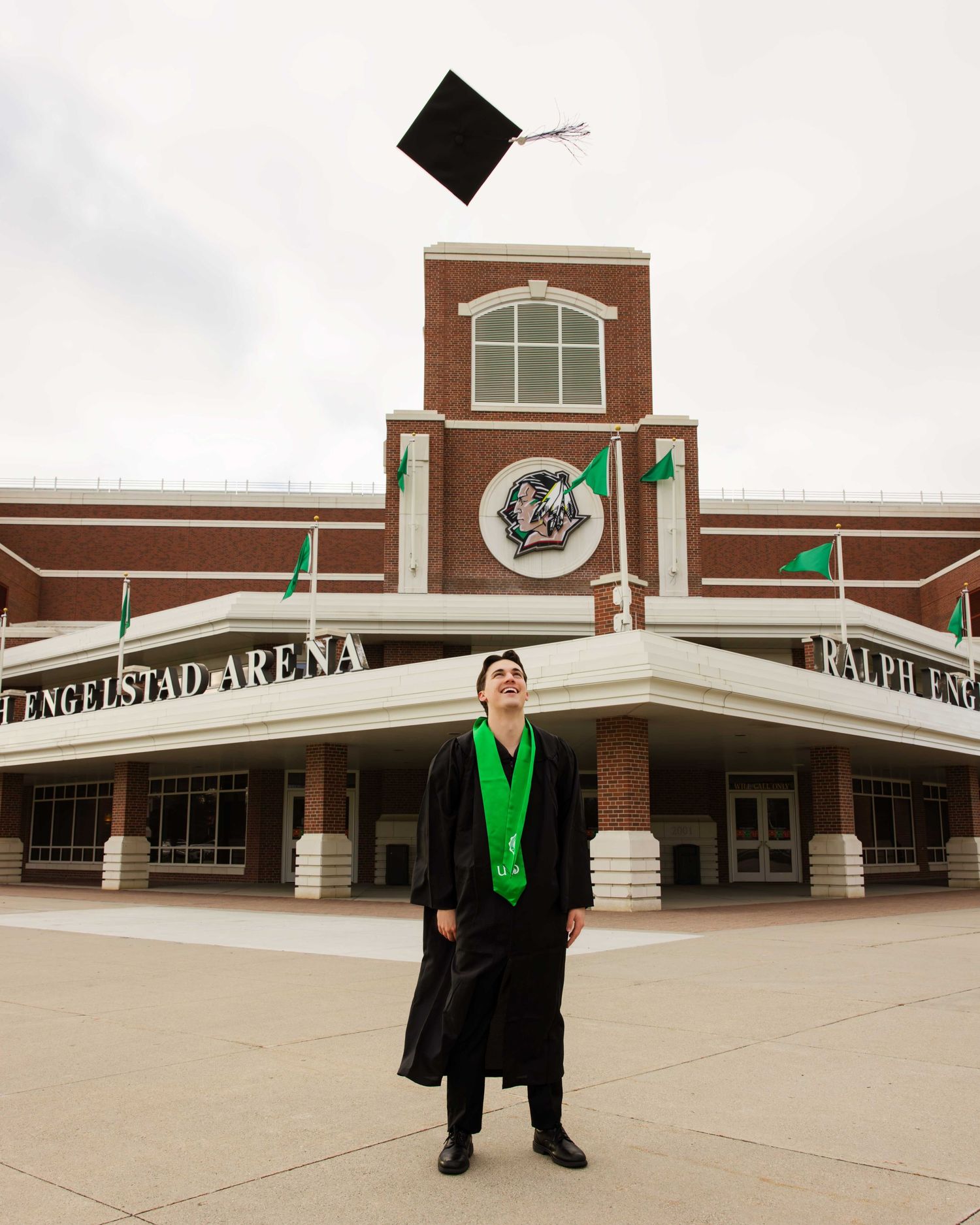 Graduate posing in black cap and gown with green stole outside brick building entrance.
