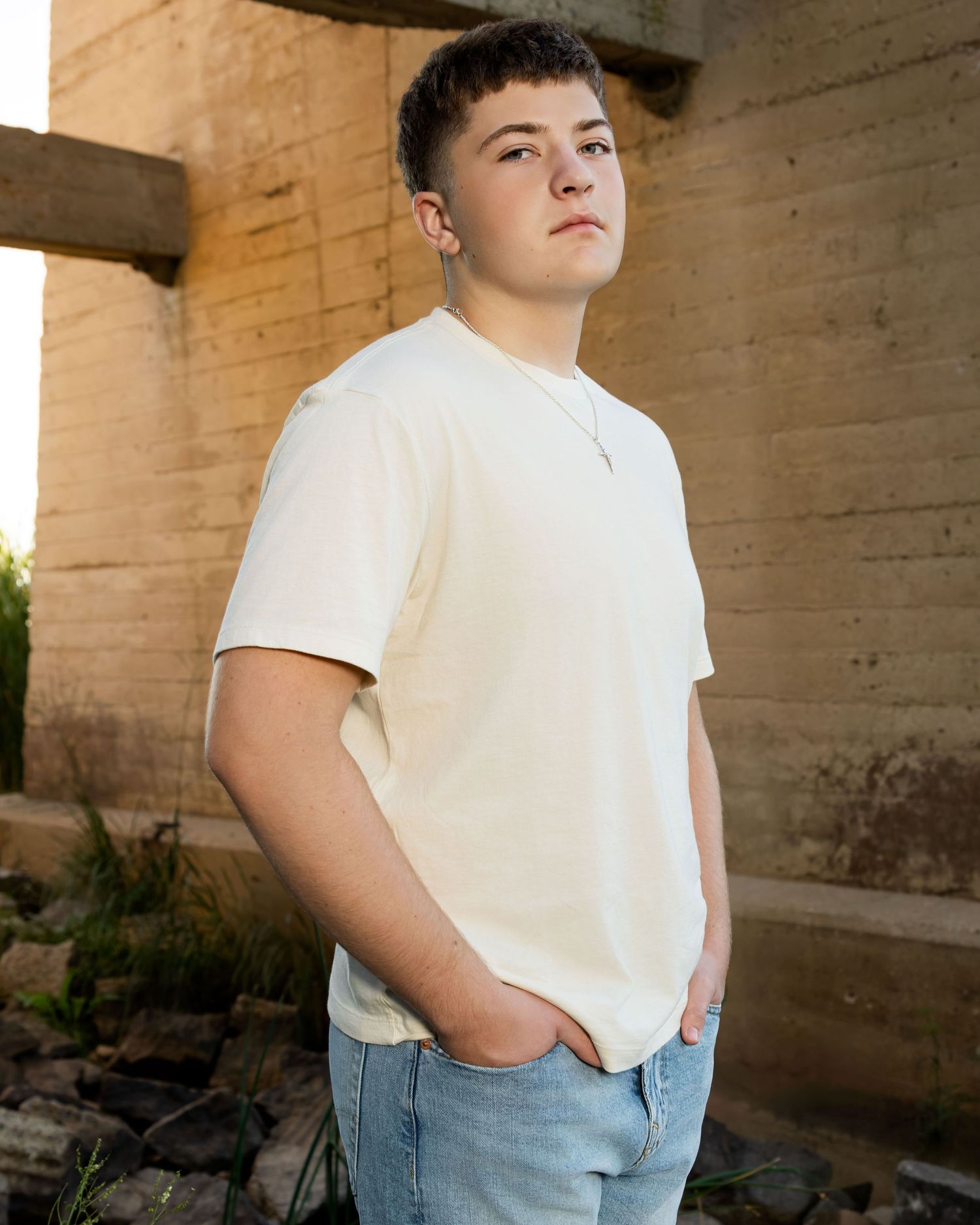 A young man in a cream t-shirt and light jeans poses with hands in pockets near a rustic stone wall outdoors.