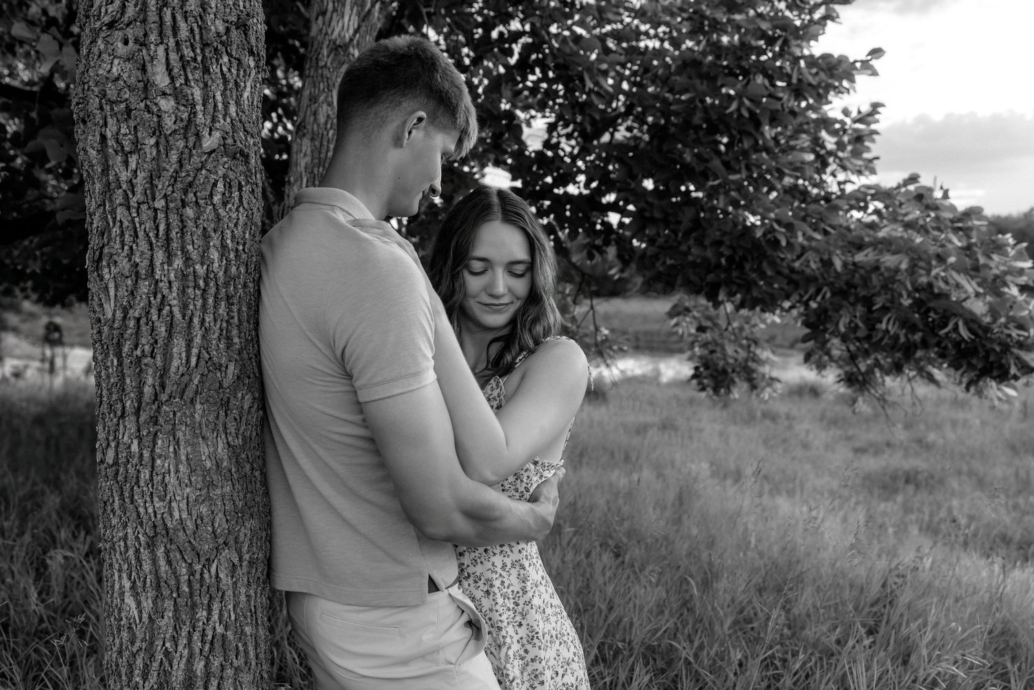 Black and white photo of a young couple embracing tenderly by a tree in a scenic outdoor field.