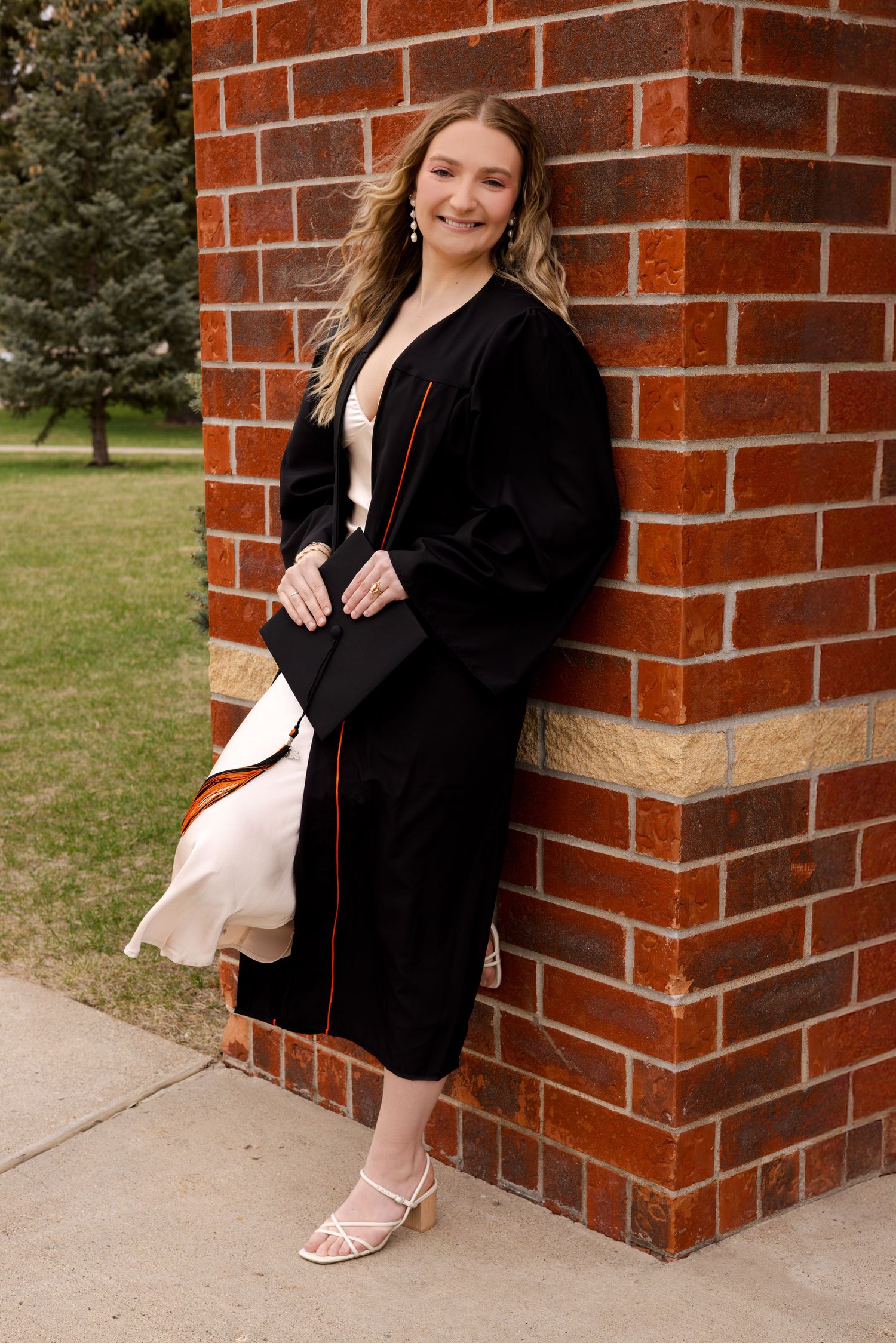 Graduate in black dress sits against brick wall during graduation photoshoot.