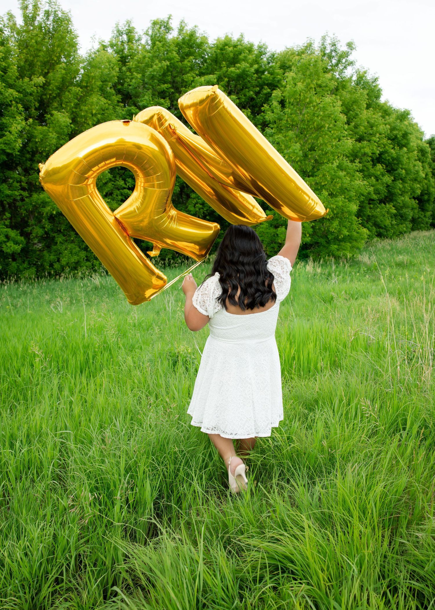 Woman in white dress holding gold RN letter balloons in a lush green field, celebrating nursing graduation.