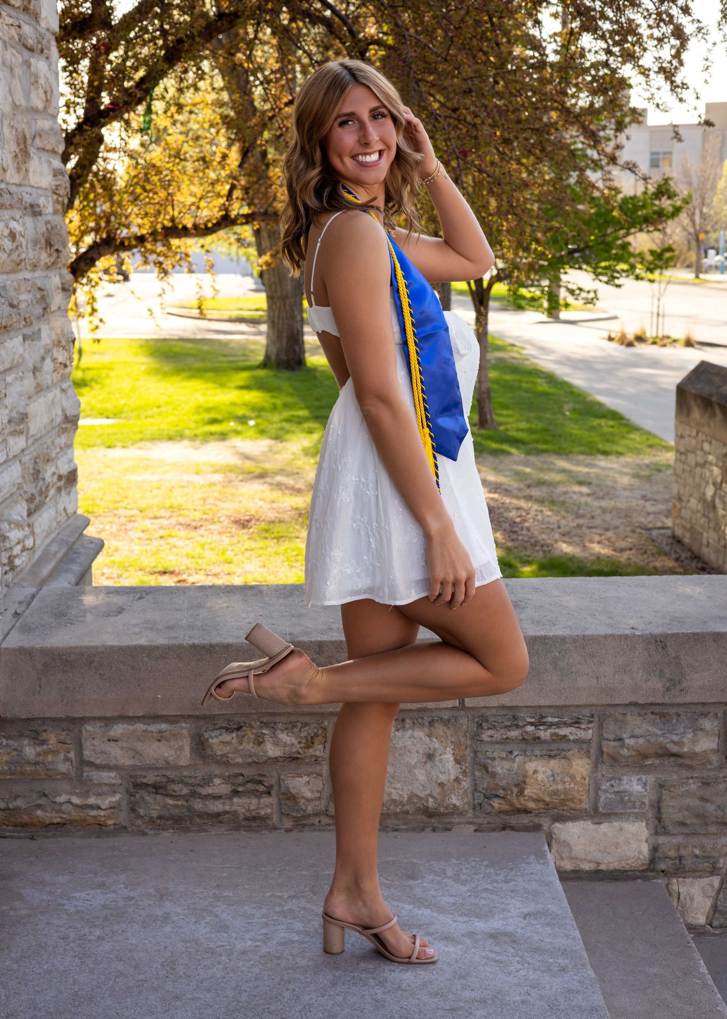 Graduate in blue dress poses on stone wall during golden hour photoshoot.