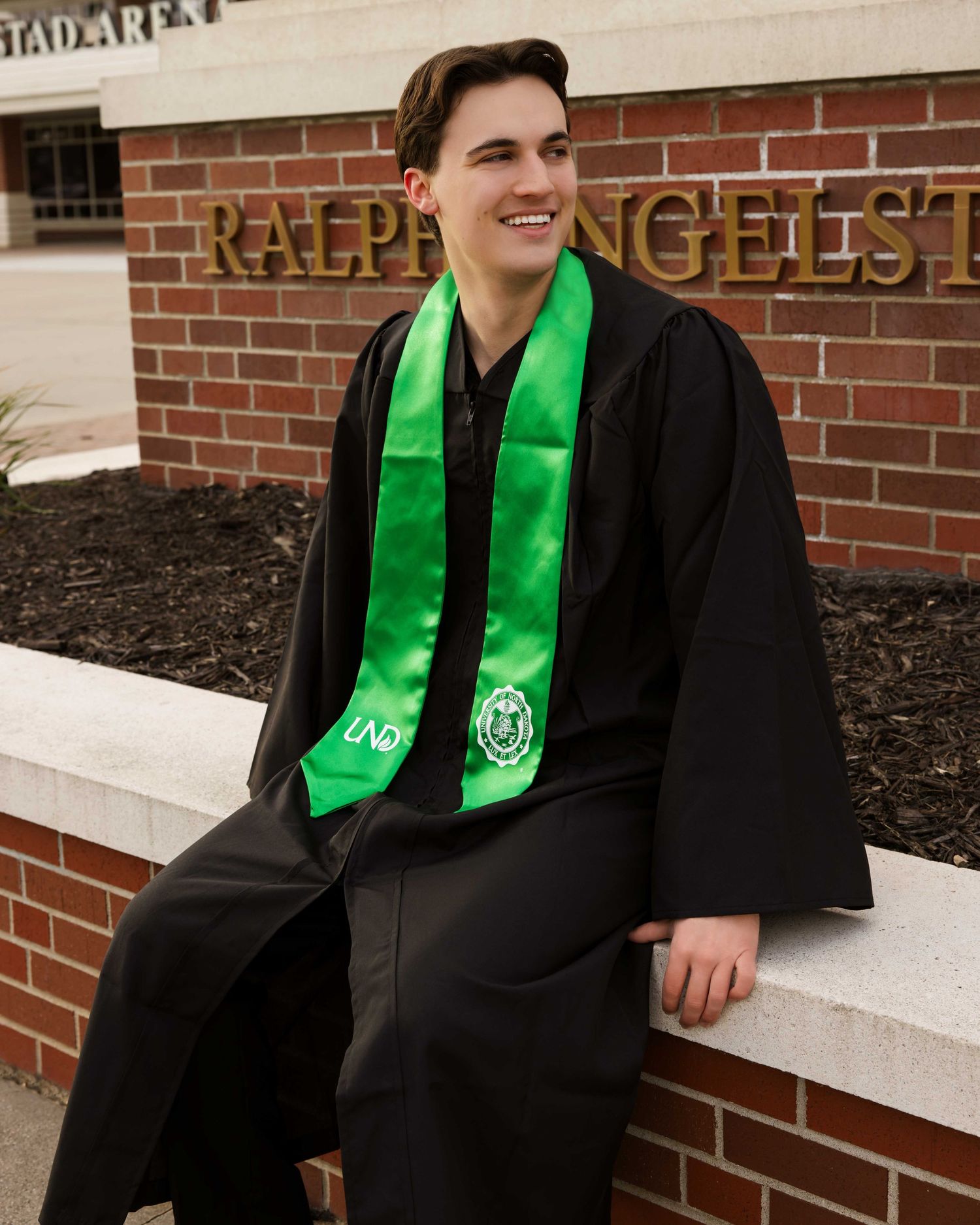 Graduate wearing black gown and bright green stole sits on brick ledge for graduation photos.