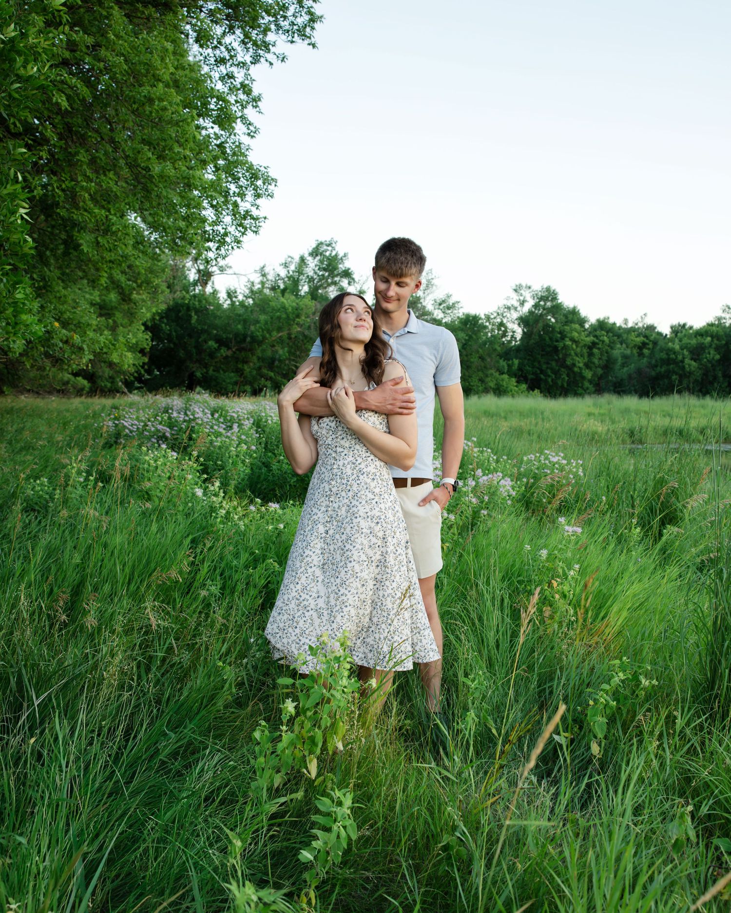 A young couple embraces romantically in a lush green meadow, the woman in a floral dress leaning back into her partner.
