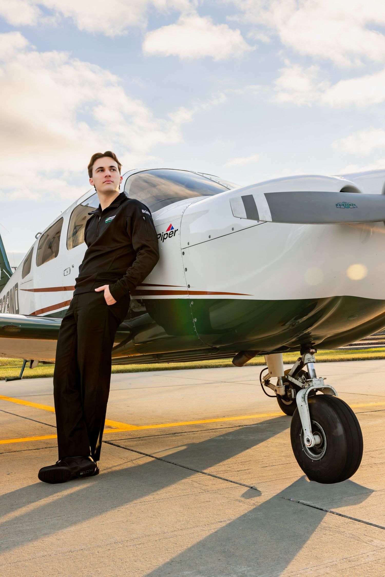 Young pilot in uniform stands confidently beside a Piper aircraft on a sunny airport tarmac.