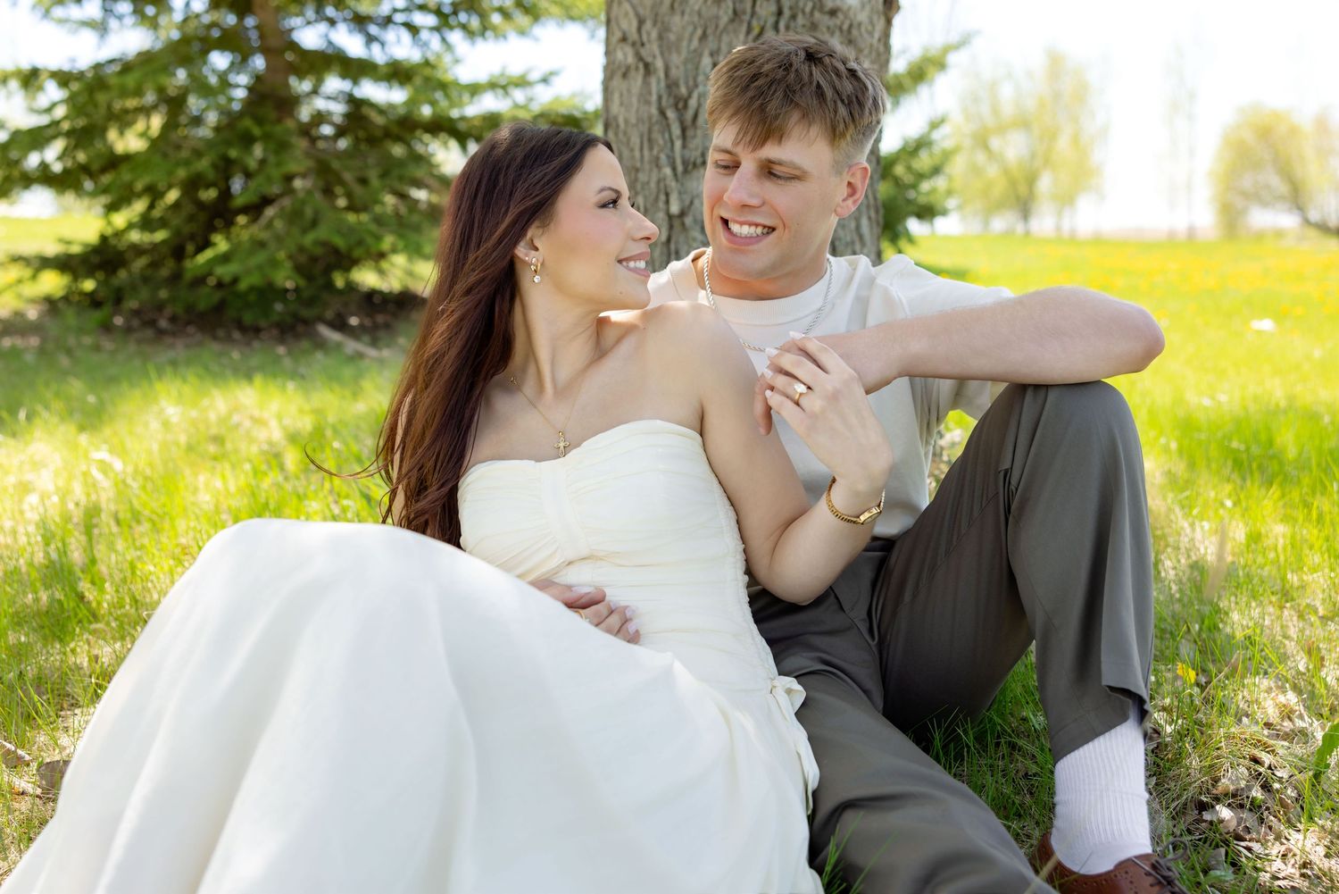A bride and groom sit together under a tree in a sunny green field, smiling and holding hands on their wedding day.
