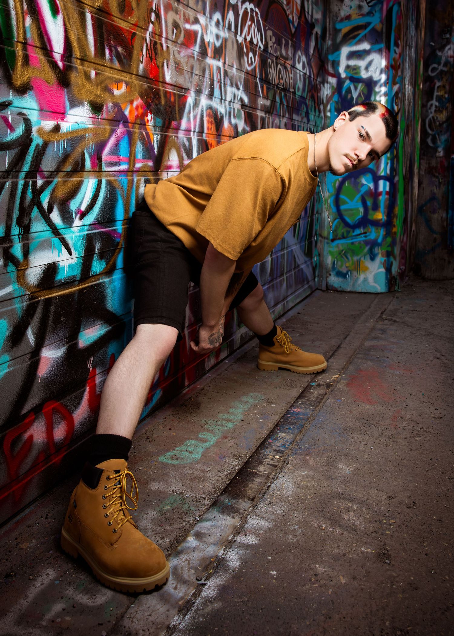 Young person in mustard tee, black shorts, and tan boots posing against colorful graffiti wall in urban alley.