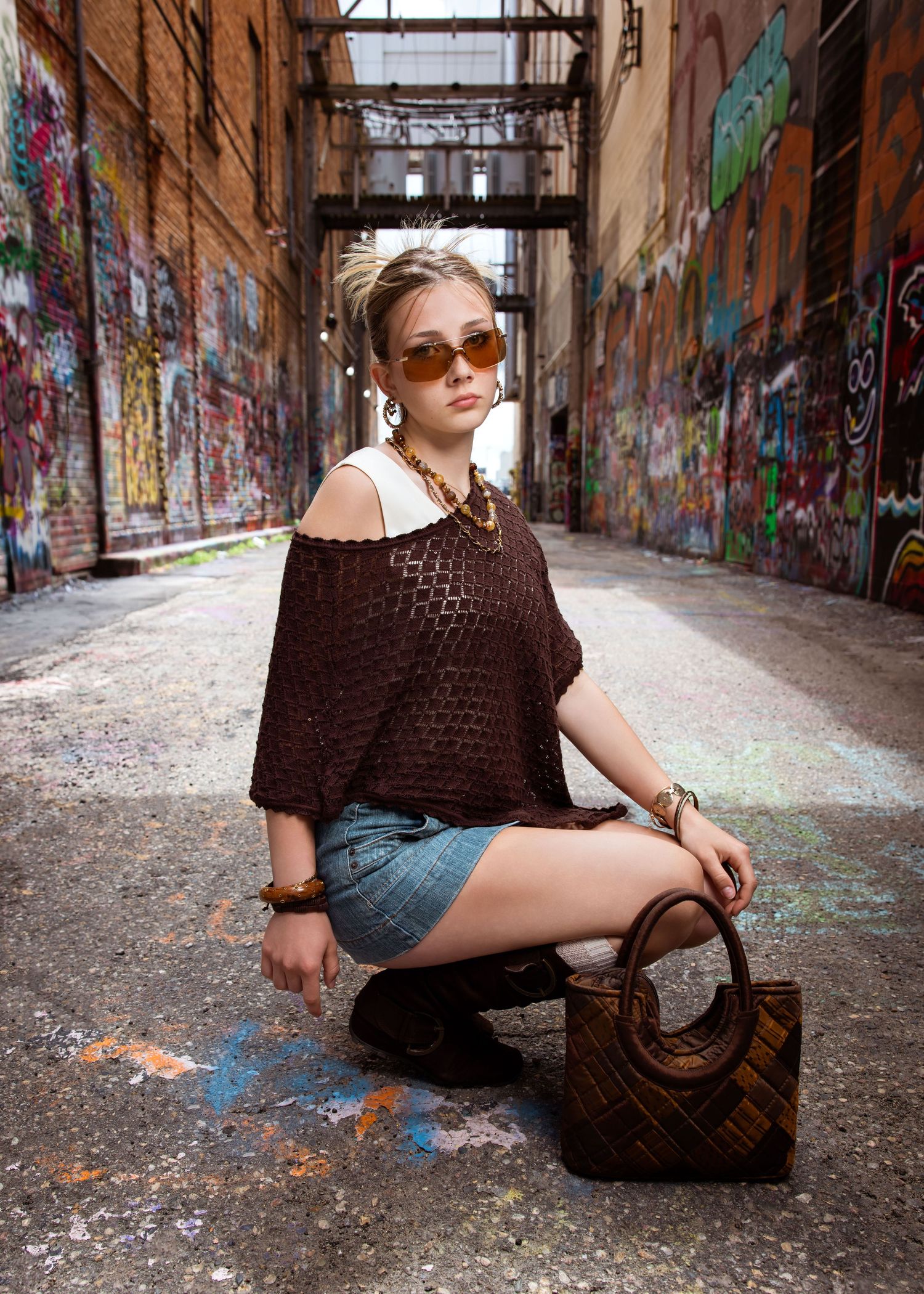 Young woman in brown mesh top, denim shorts, sunglasses, posing in graffiti-covered urban alley with woven handbag.