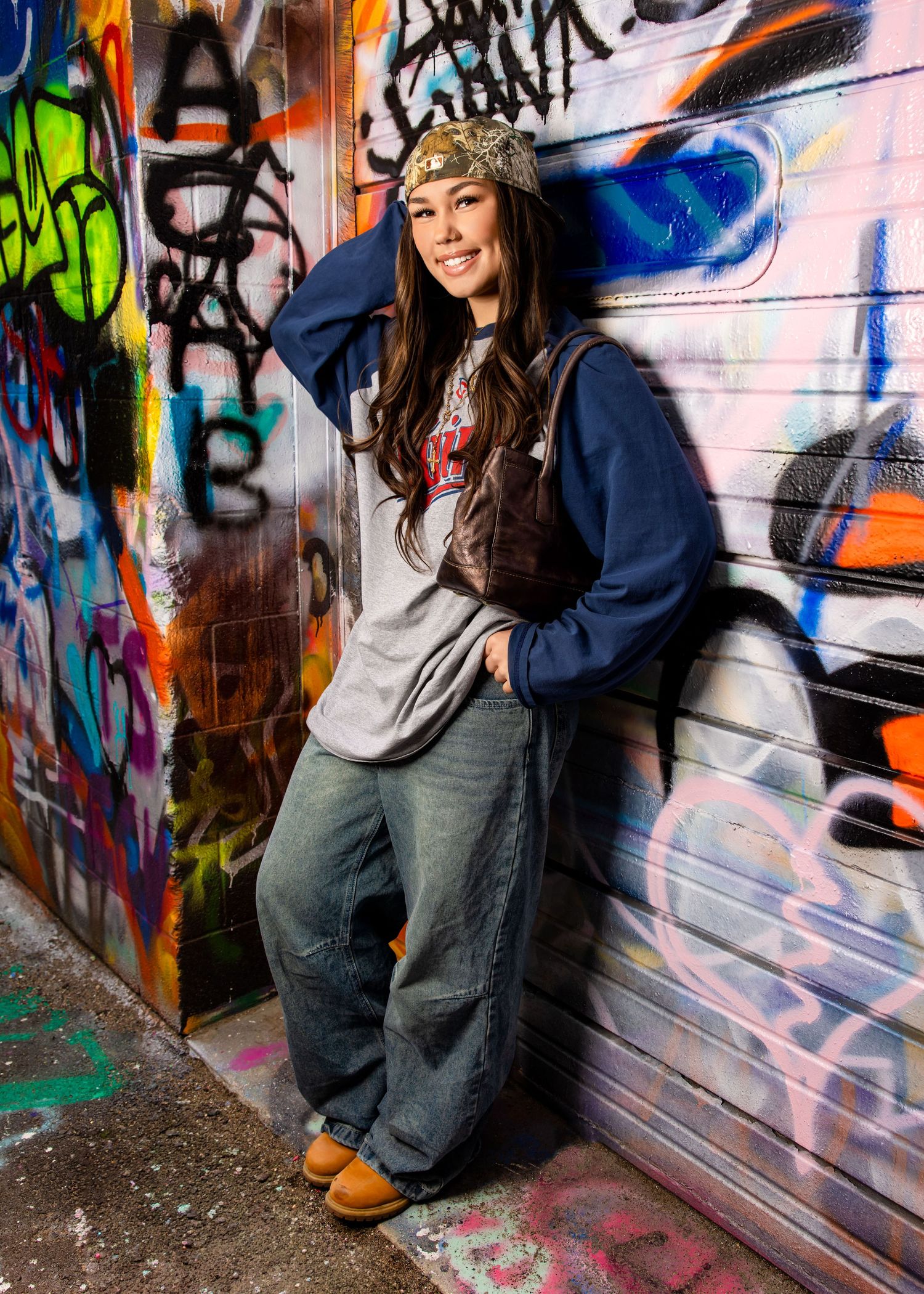 Smiling woman in baggy jeans, navy hoodie, and bandana leaning against vibrant graffiti-covered wall outdoors.