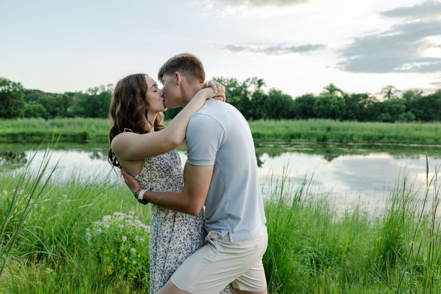 Couple sharing a romantic kiss by a serene pond surrounded by lush green grass and trees at dusk.