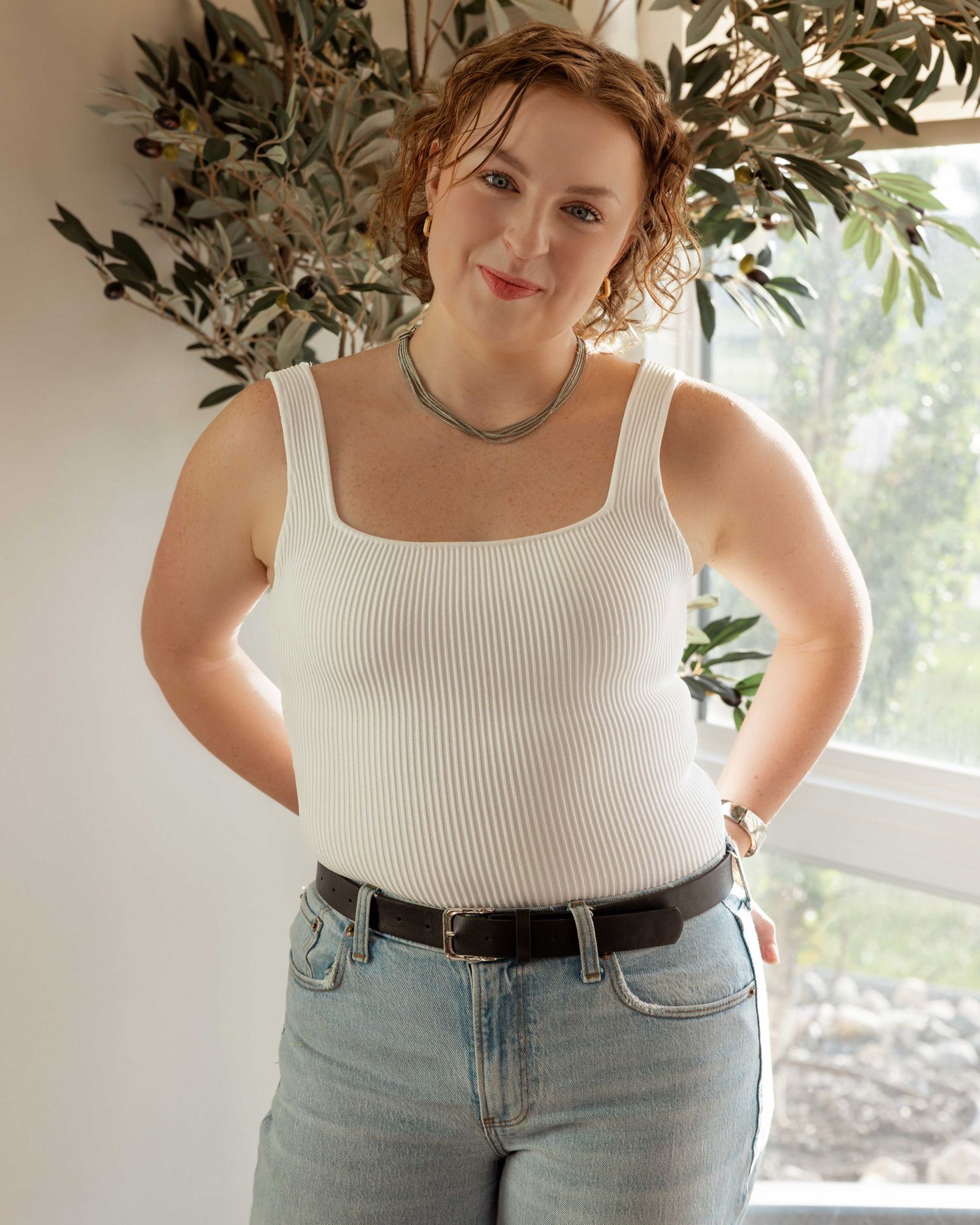 Person in white tank top and jeans posing casually by window with plants.