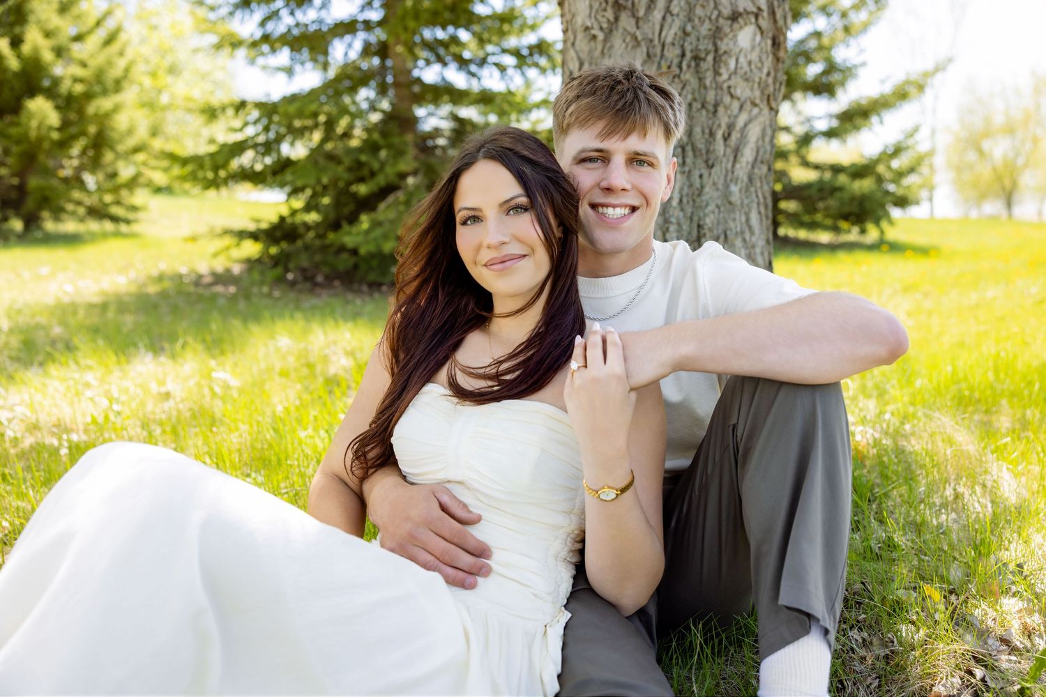 Happy couple sitting against a tree in a sunny green park, woman in white dress, man in gray pants, smiling outdoors.