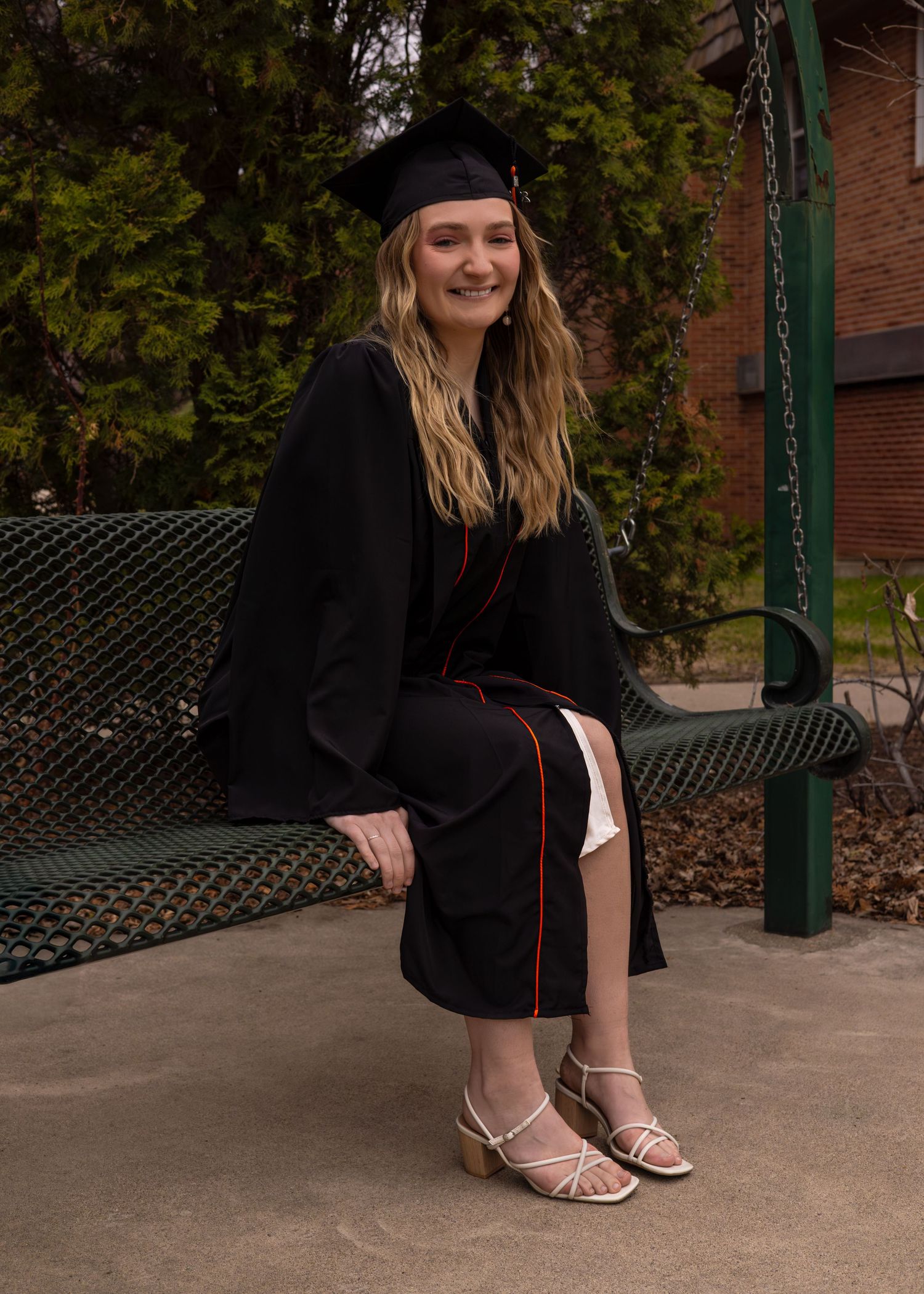 Graduate in black gown poses on concrete steps with autumn trees in background.