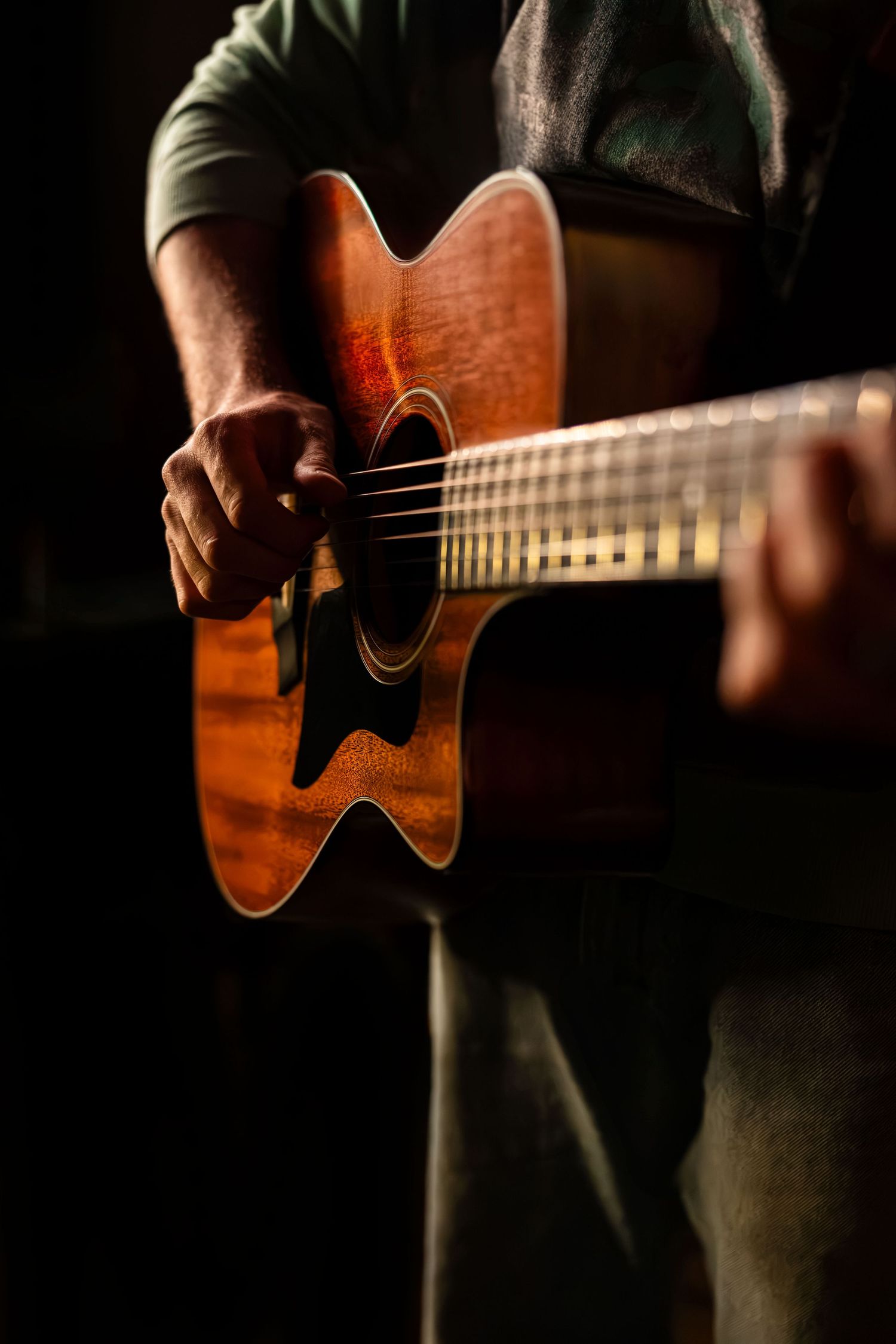 A musician plays an acoustic guitar in dramatic low-key lighting, highlighting the warm wood tones of the instrument.
