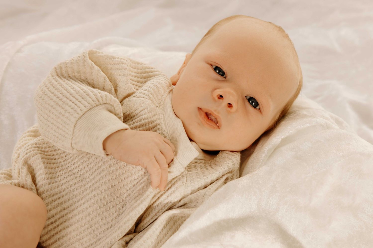 Sweet newborn baby in cream-colored outfit lying on white bedding.
