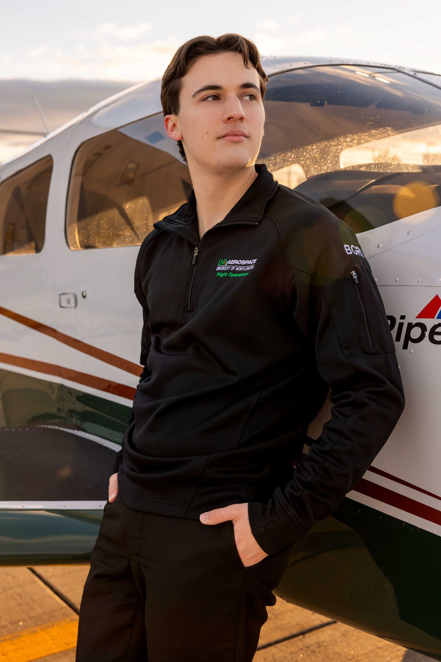 Professional pilot poses in black uniform beside small aircraft at sunset.