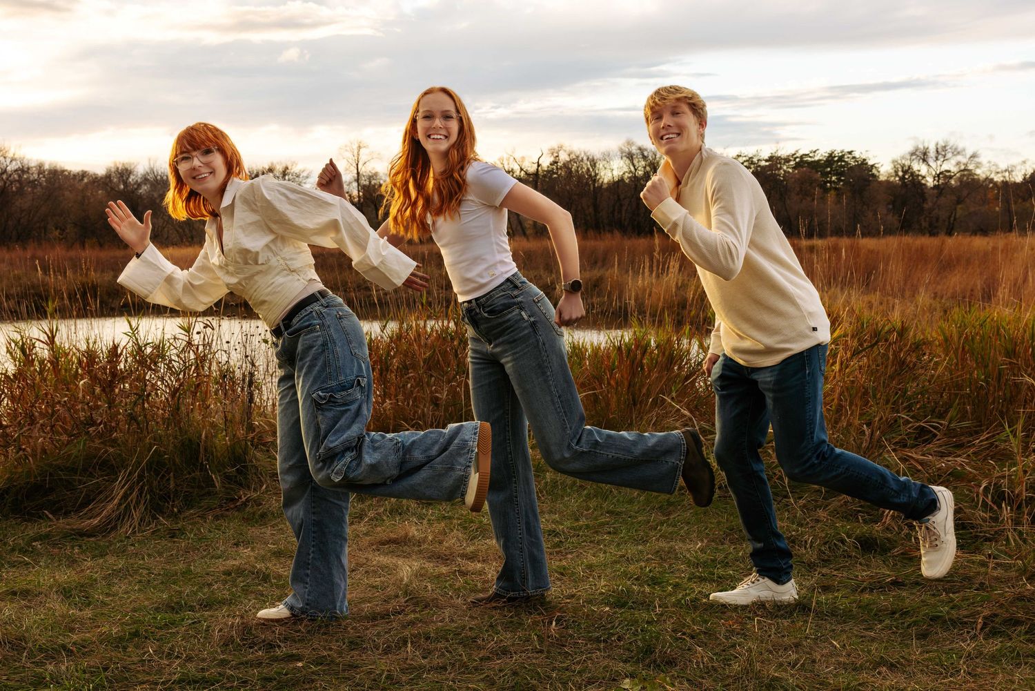 Three friends in casual wear dance playfully in autumn field at sunset.