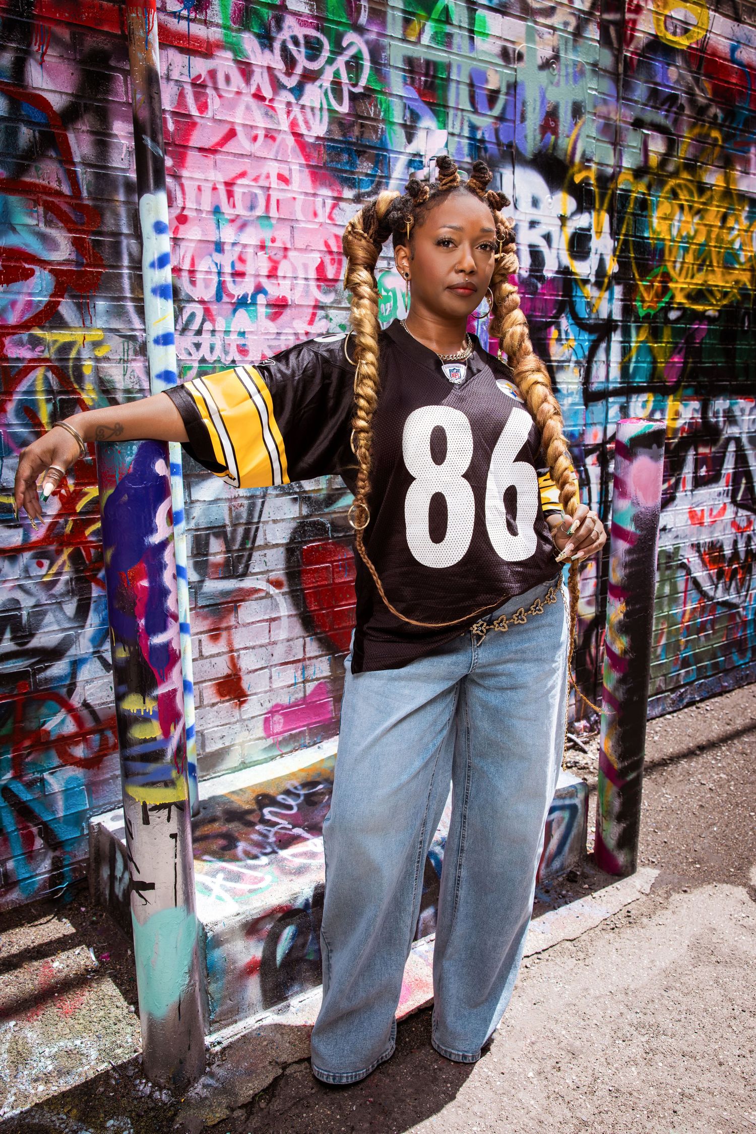 Woman in NFL jersey 86 with braids posing against vibrant graffiti wall in urban setting.