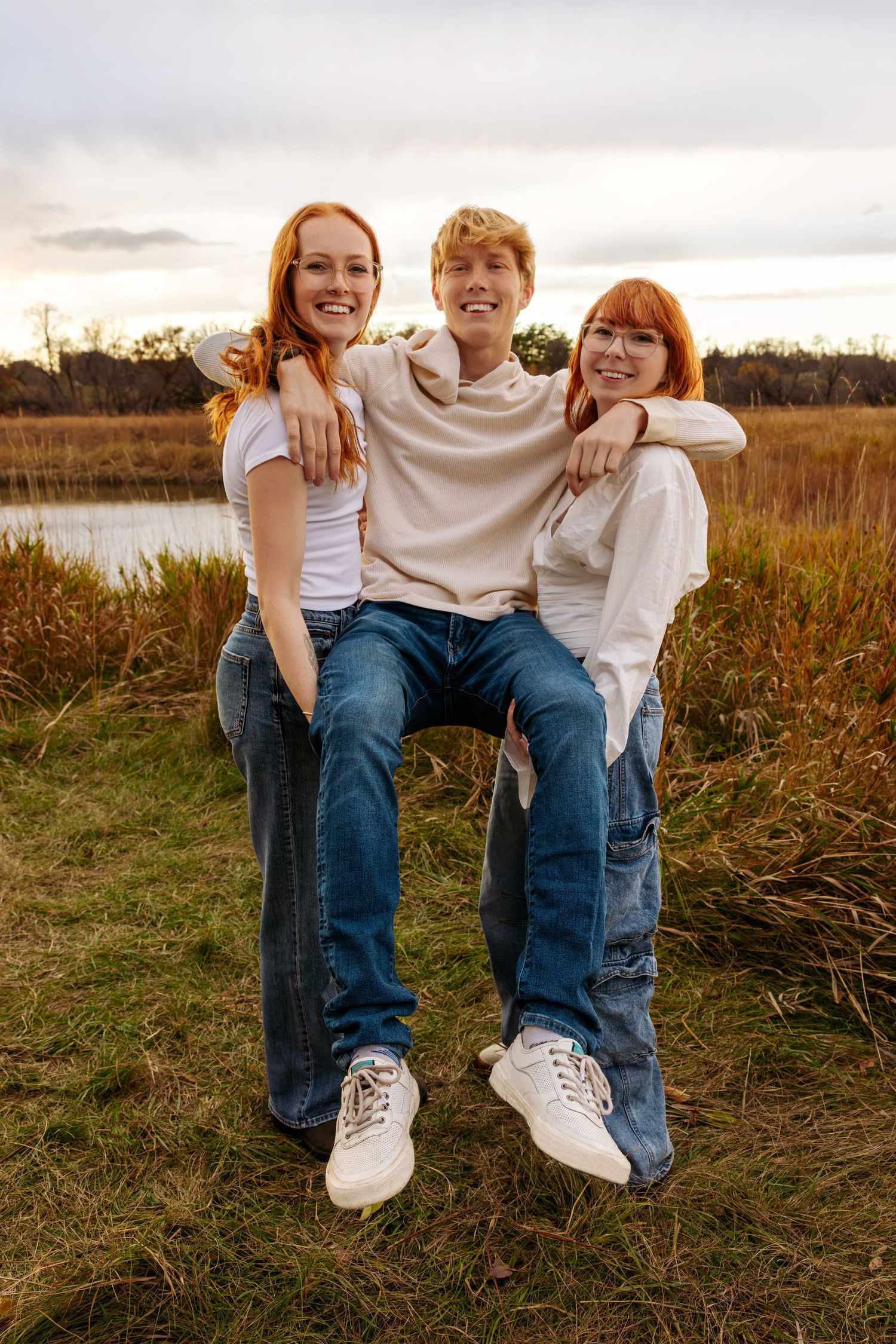 Friends in casual denim and white outfits sit together laughing in a grassy field at sunset.