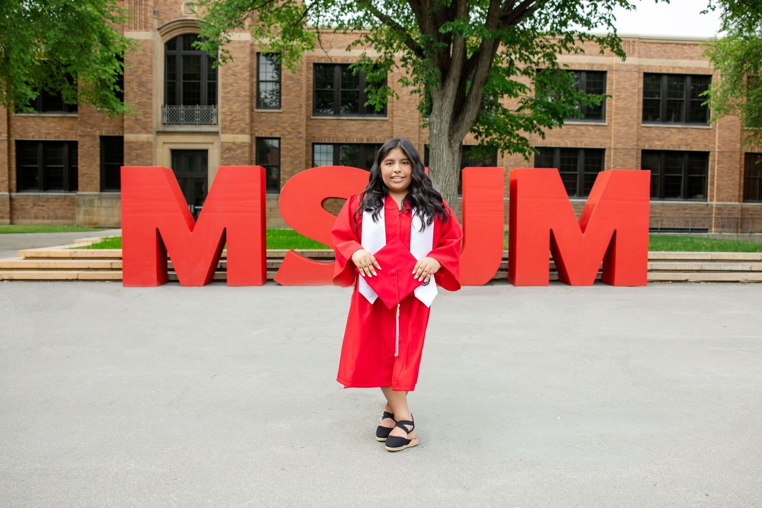 Graduate in red cap and gown posing in front of large red MSJM letters on a university campus.