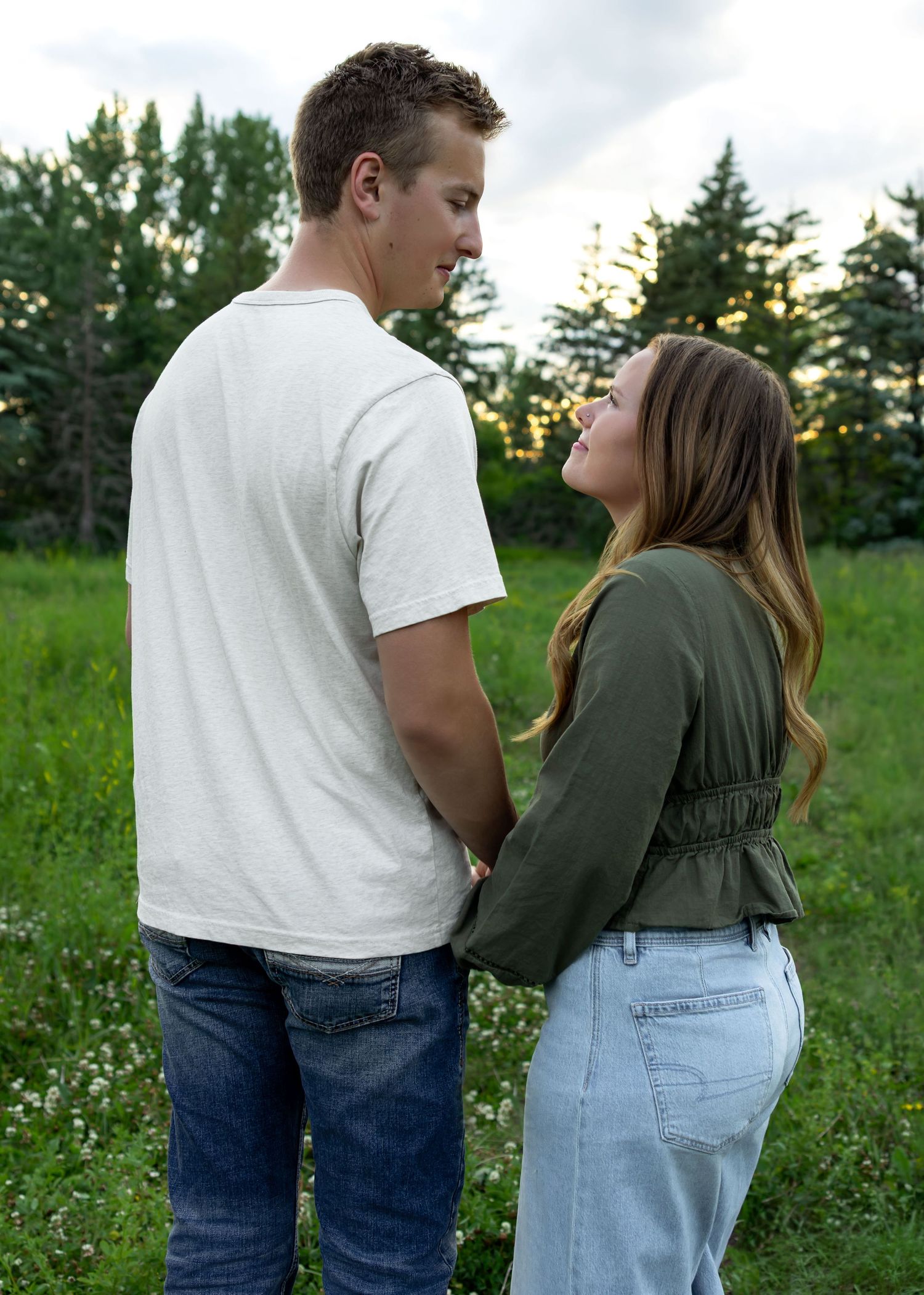 A young couple holding hands and gazing at each other in a lush green meadow at dusk.