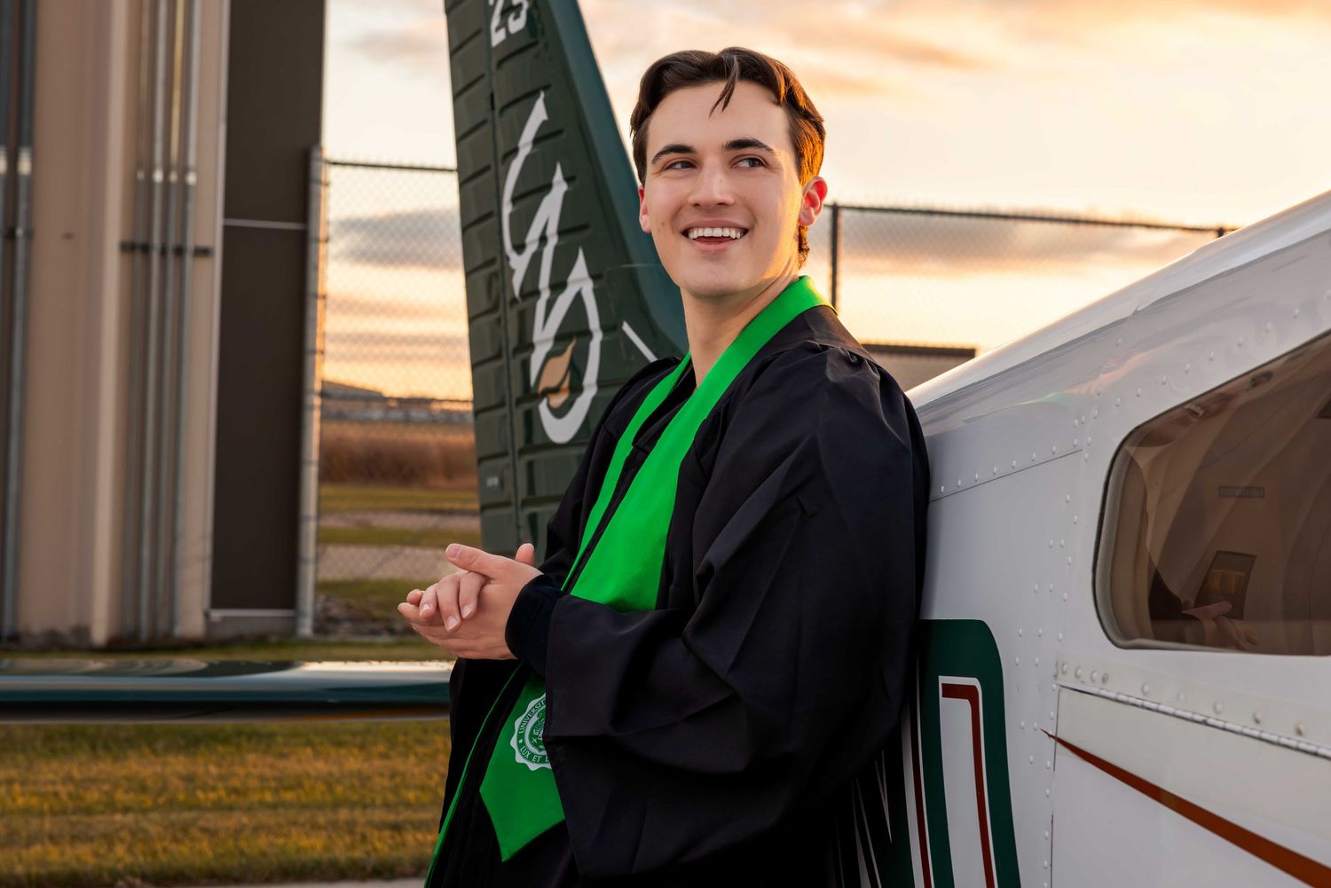 Graduate in black gown with green stole poses next to small aircraft at sunset.