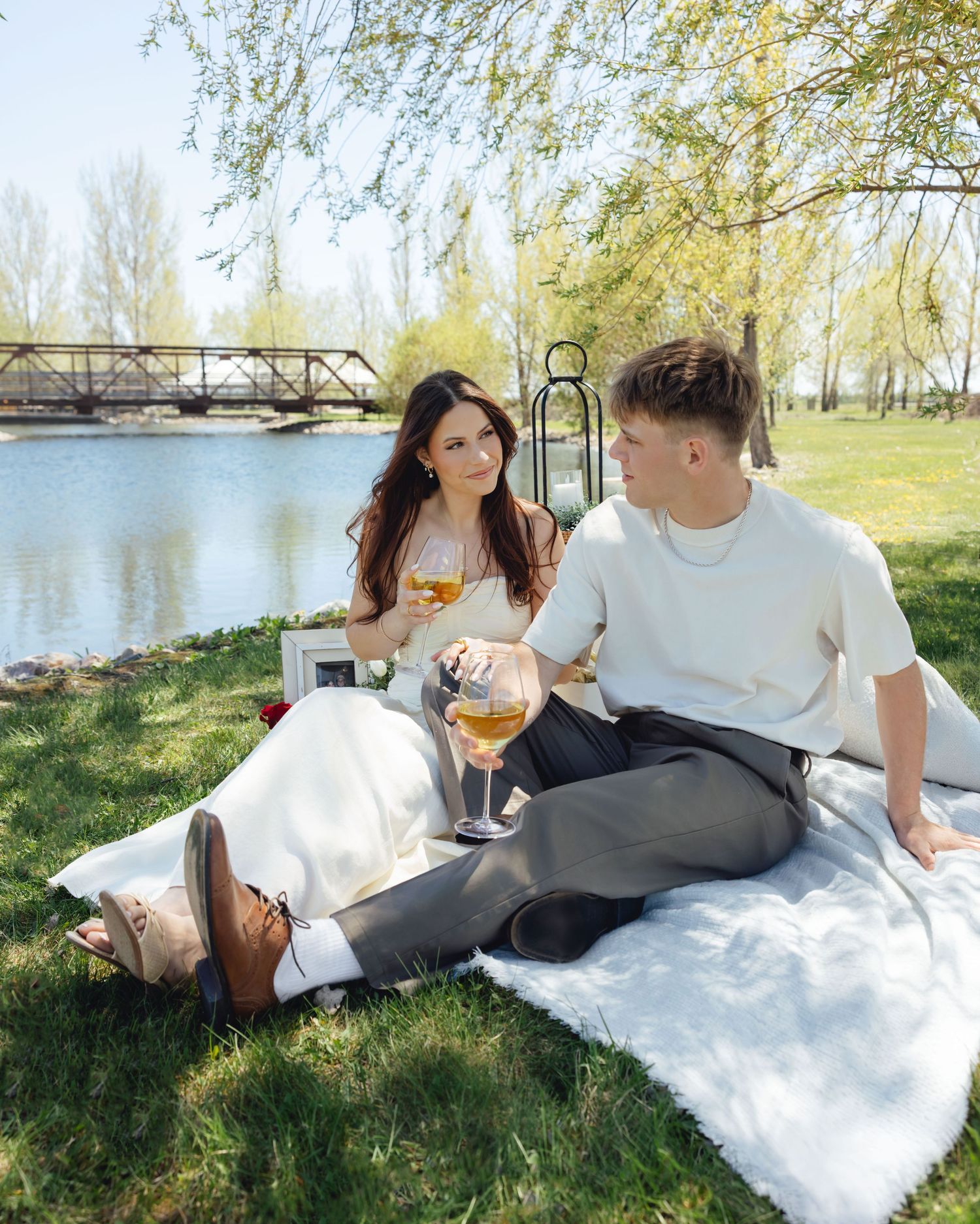 Couple enjoying a romantic outdoor picnic by a river, holding wine glasses on a sunny spring day.