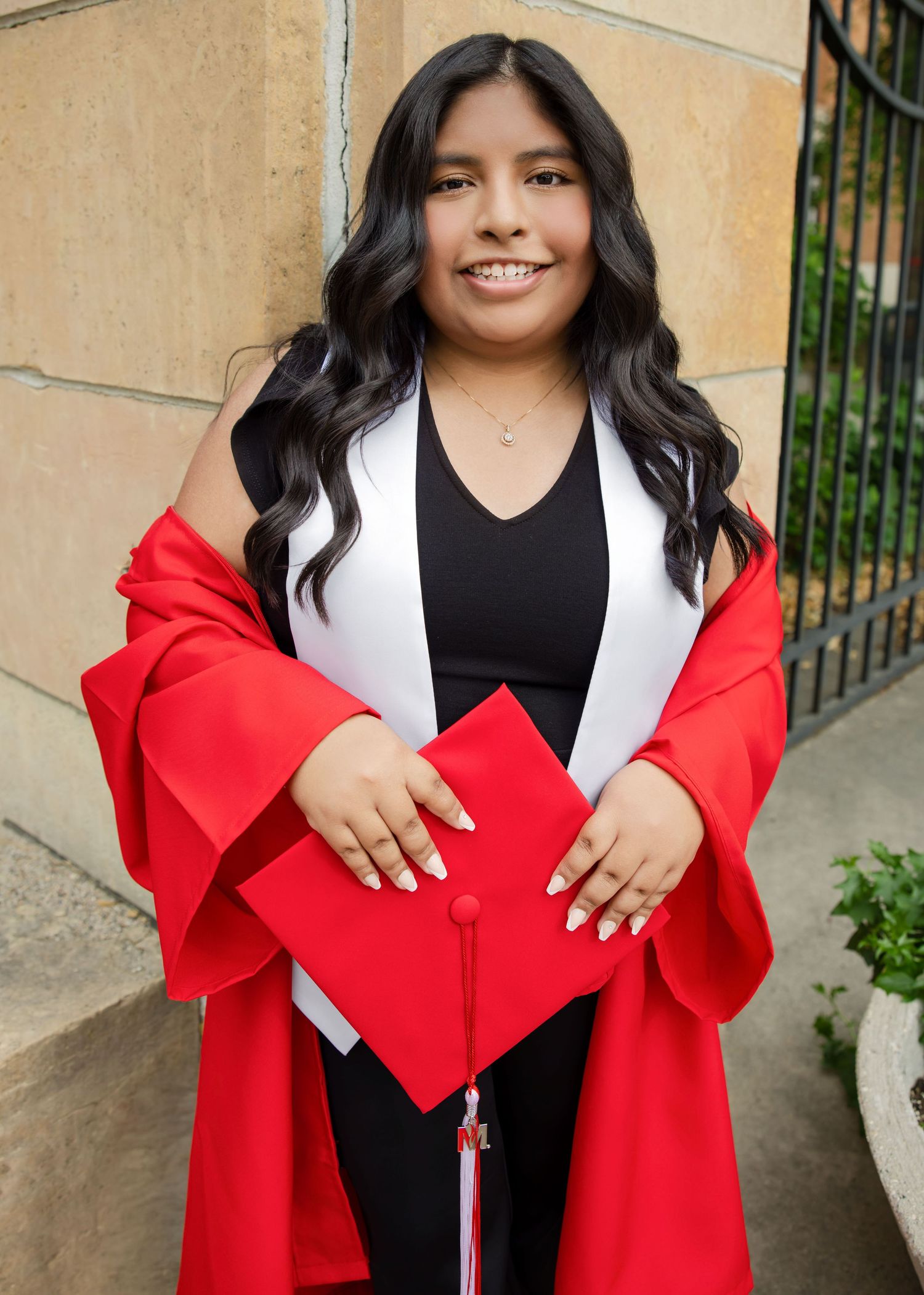 Young woman in red graduation gown and cap smiling proudly outdoors on her graduation day.