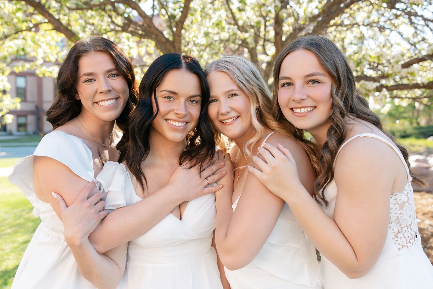 Four bridesmaids in white dresses smiling and posing together outdoors under trees at a wedding celebration.