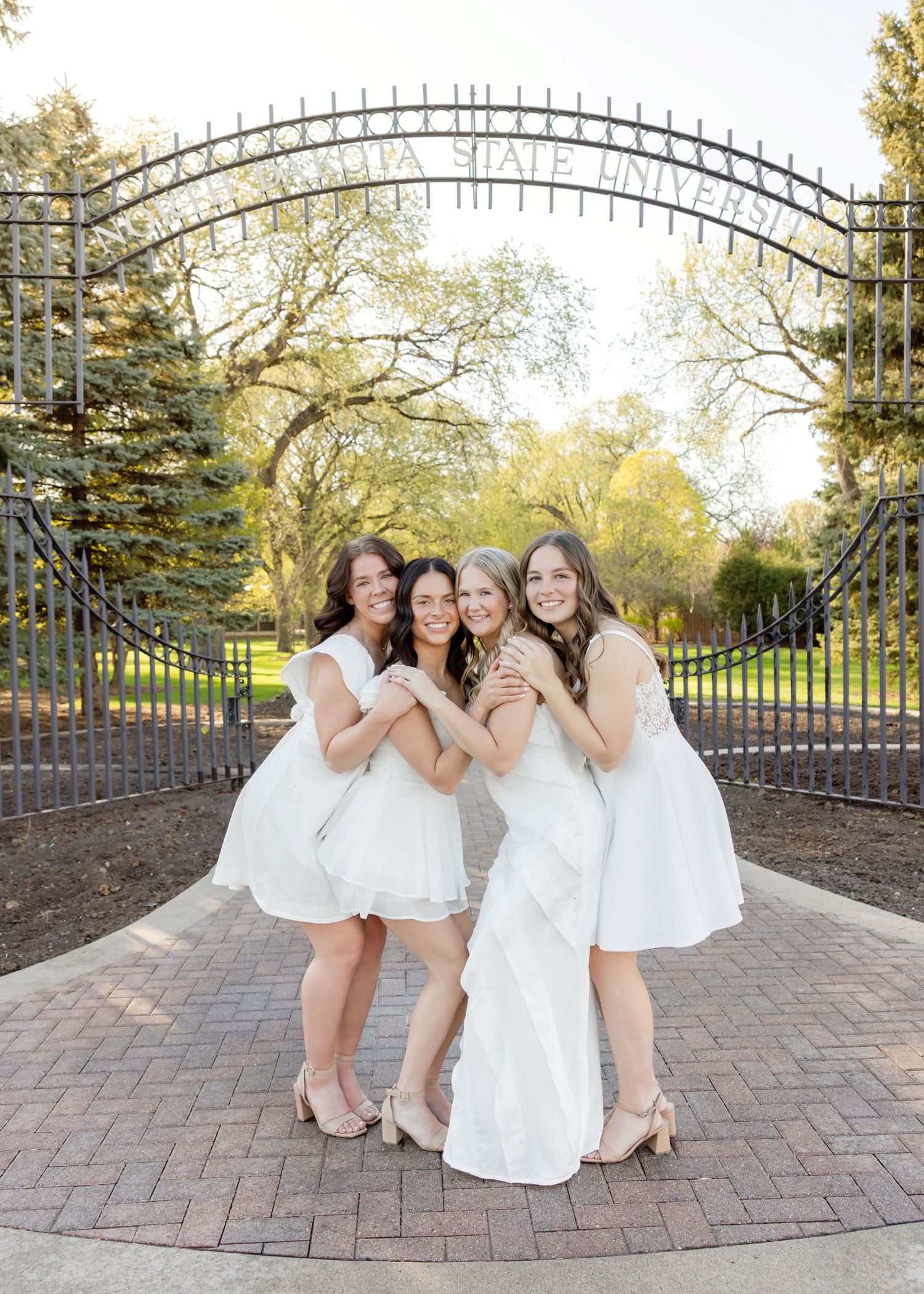 Four women in white dresses pose together at a university gate entrance on a sunny day.