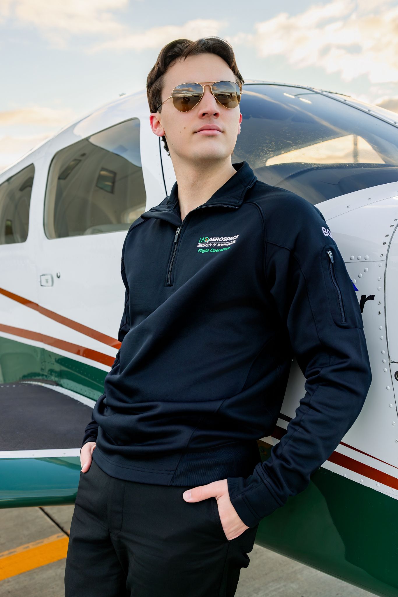 Young male pilot in aviator sunglasses and navy flight academy jacket stands confidently beside a small aircraft outdoors.