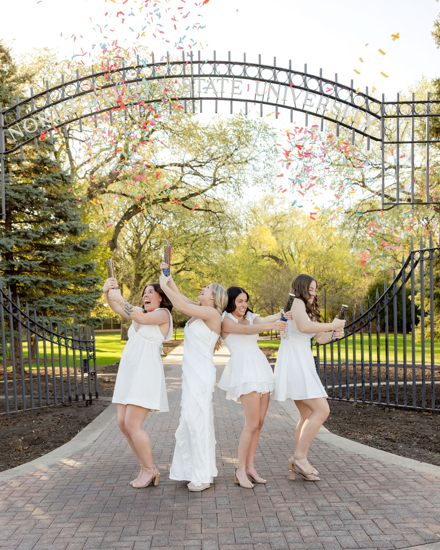 Bride and bridesmaids in white dresses celebrate with confetti at ornate university gates on a sunny day.