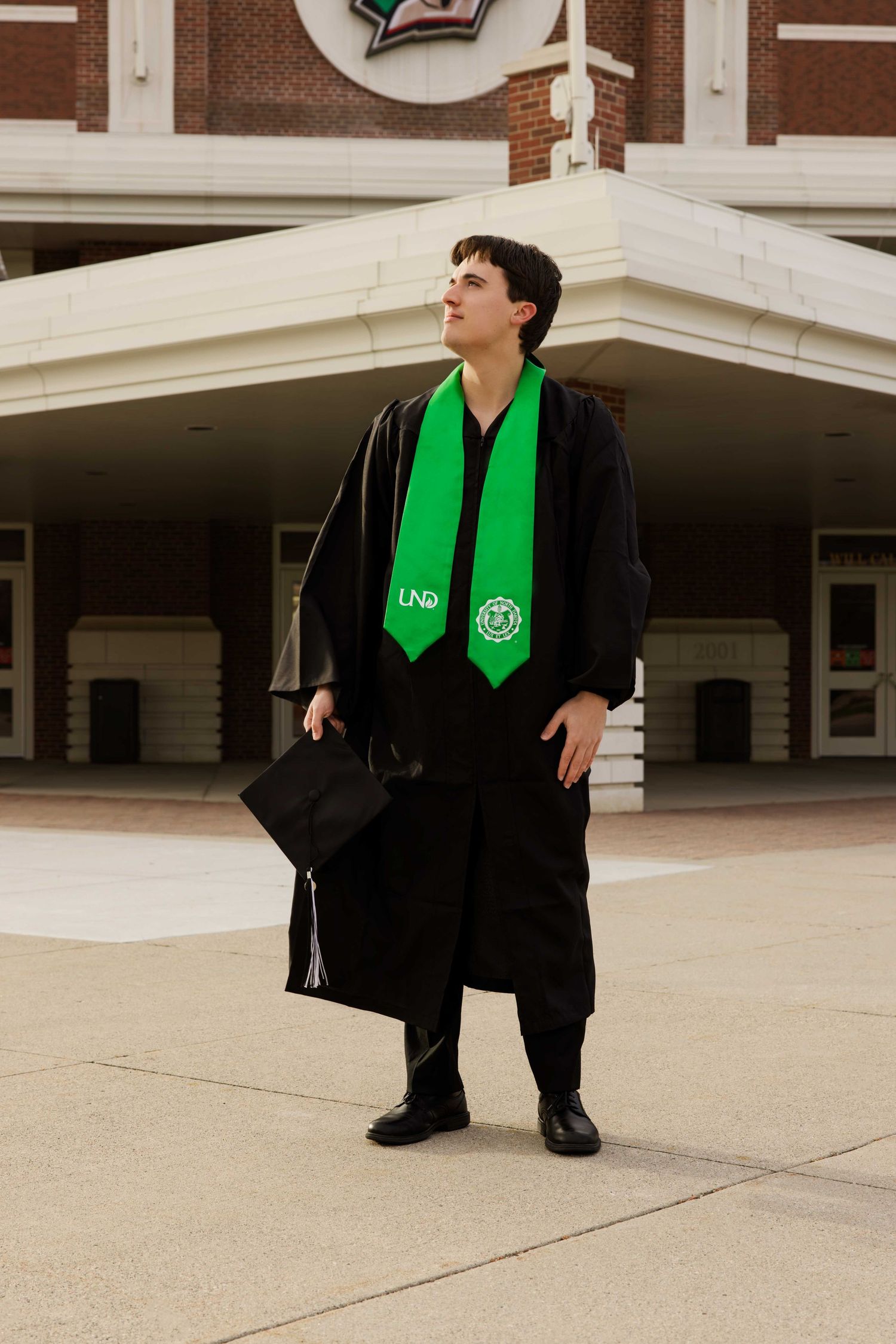 Graduate in black graduation attire with bright green stole standing in front of arena entrance.