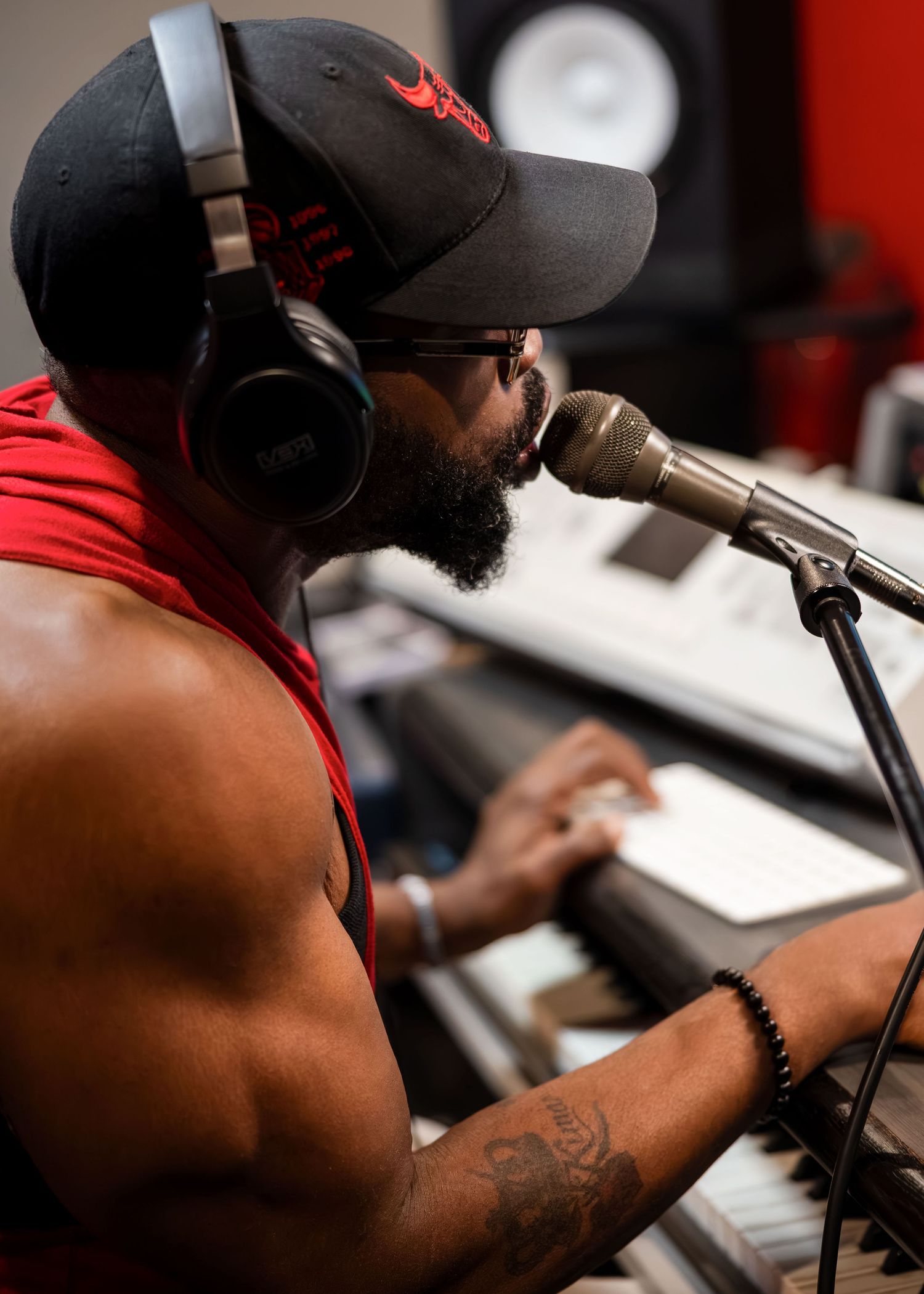 A bearded Black musician wearing headphones and a cap records vocals into a studio microphone while playing keyboard.