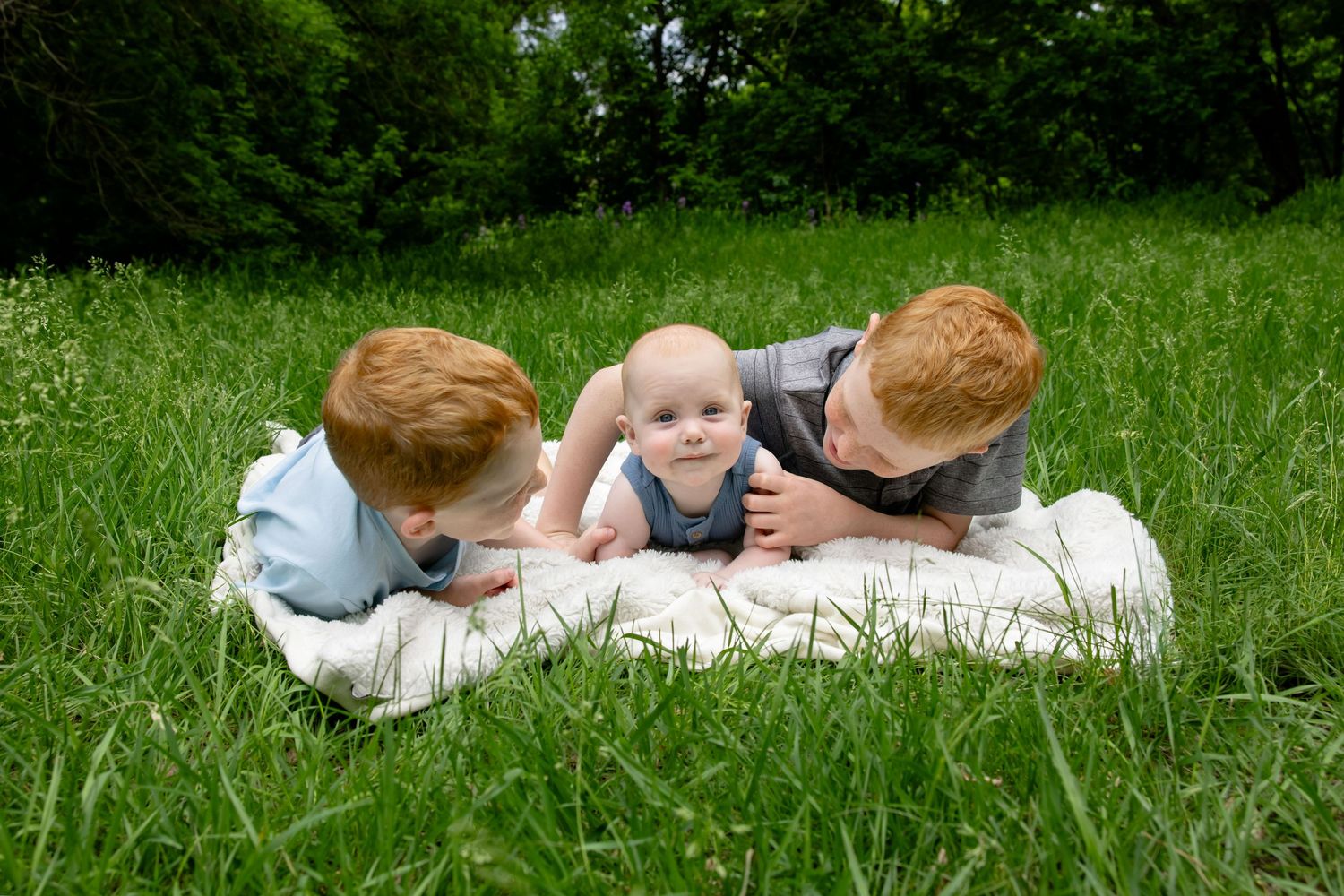 Two redhead boys lying in green grass with a baby sibling on a white blanket outdoors.