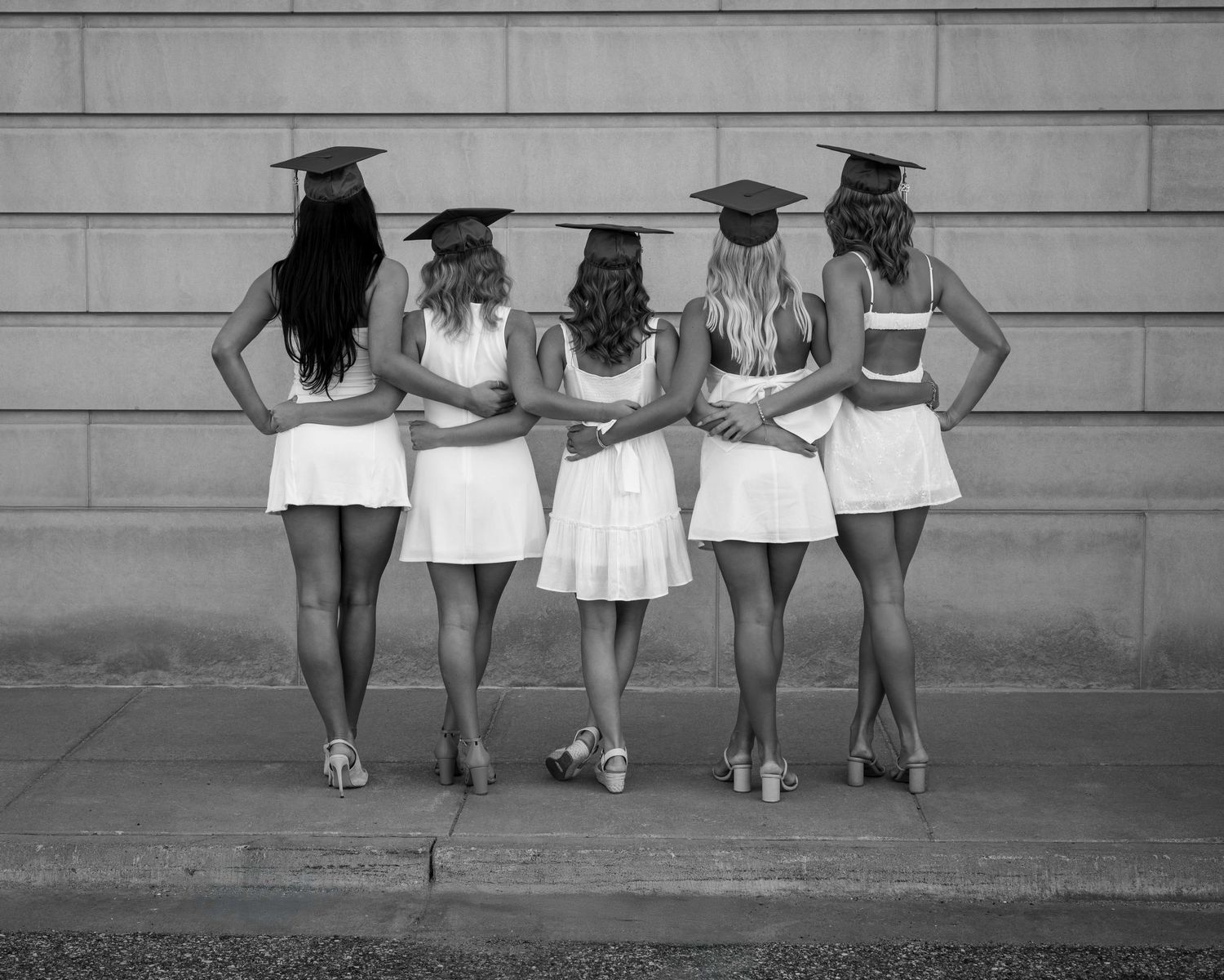 Group wearing white dresses and graduation caps posing against a wall.