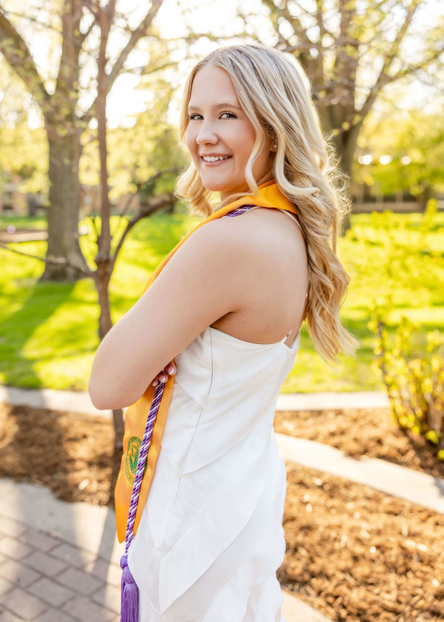 Smiling blonde woman in white dress wearing orange and purple graduation stole outdoors on a sunny day.
