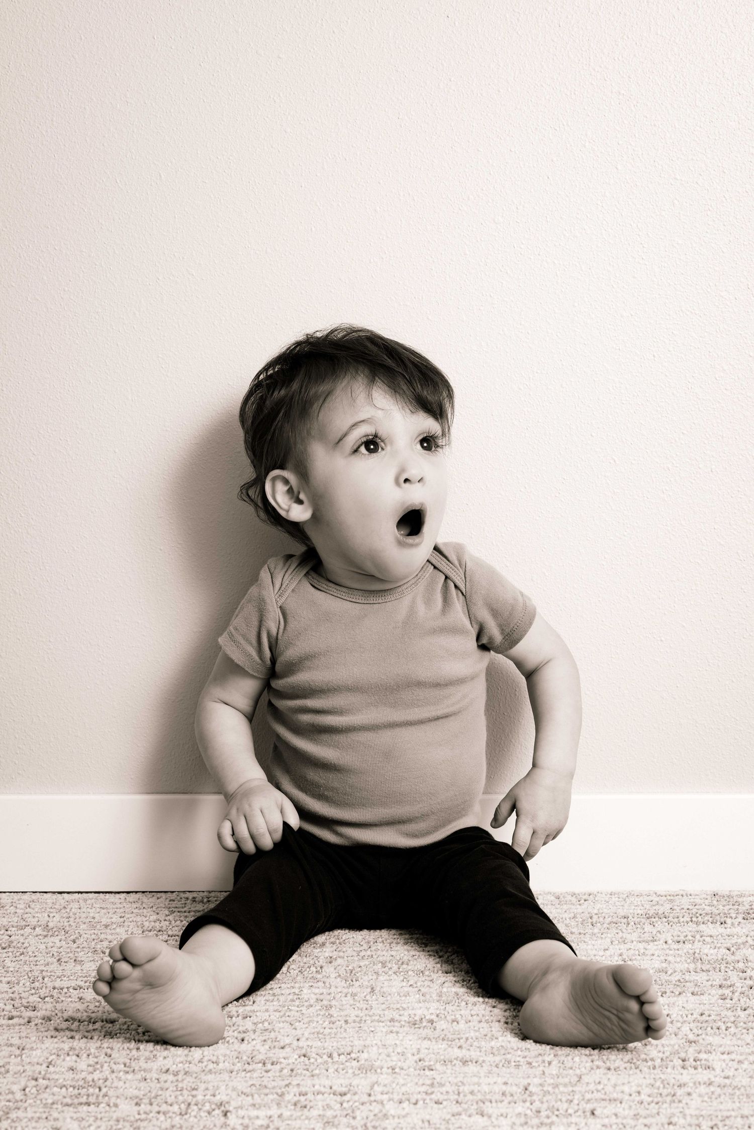 A toddler sits on carpet wearing a tan shirt with an expression of surprise and wonder.