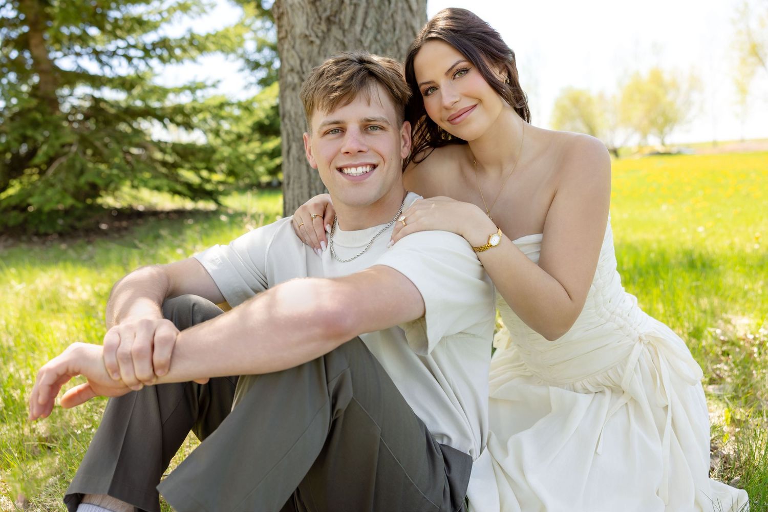 Happy newlywed couple sitting together under a tree in a sunny green field, bride in white gown and groom smiling.