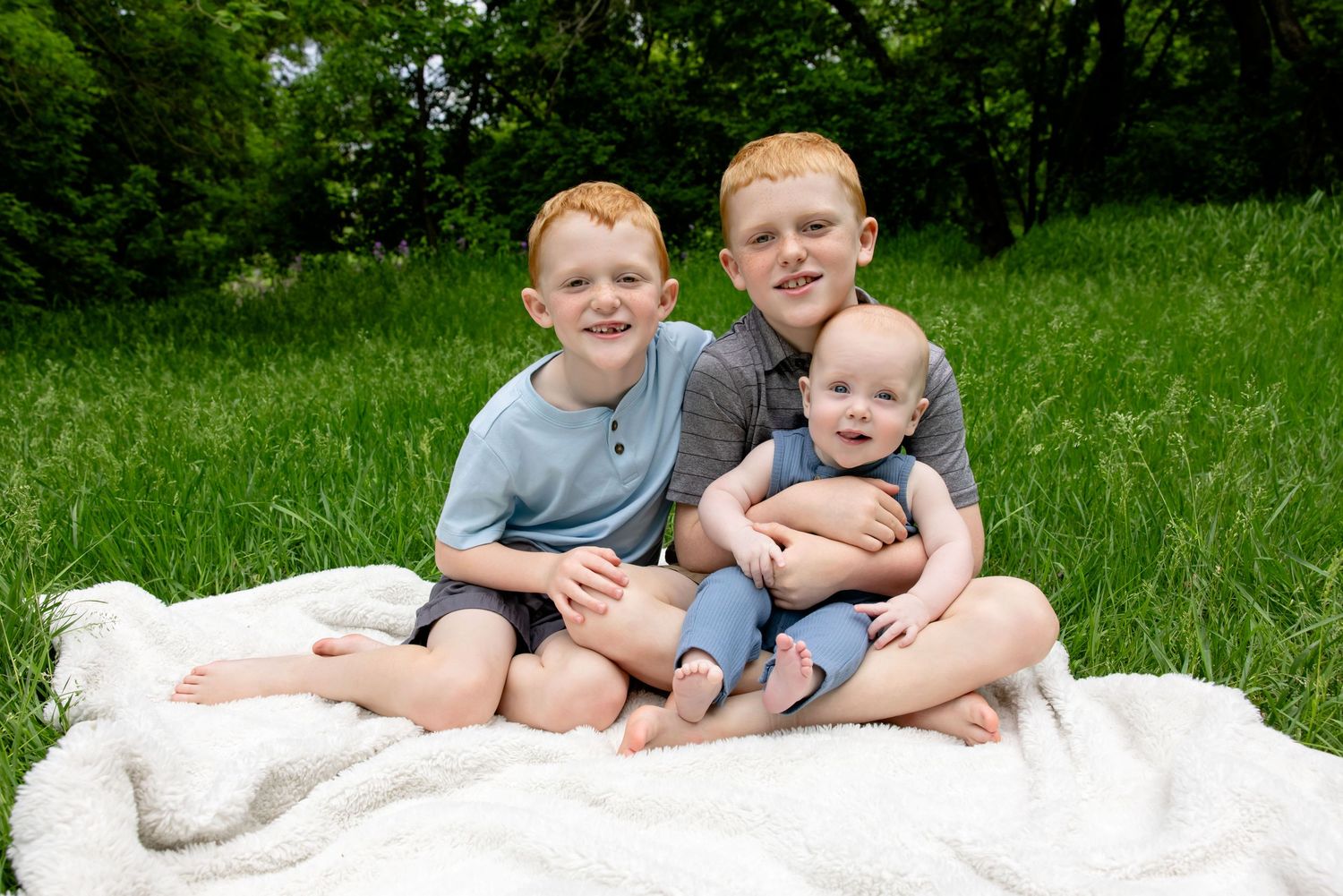 Three red-haired brothers sit together on a white blanket in a green meadow, smiling for a family portrait.