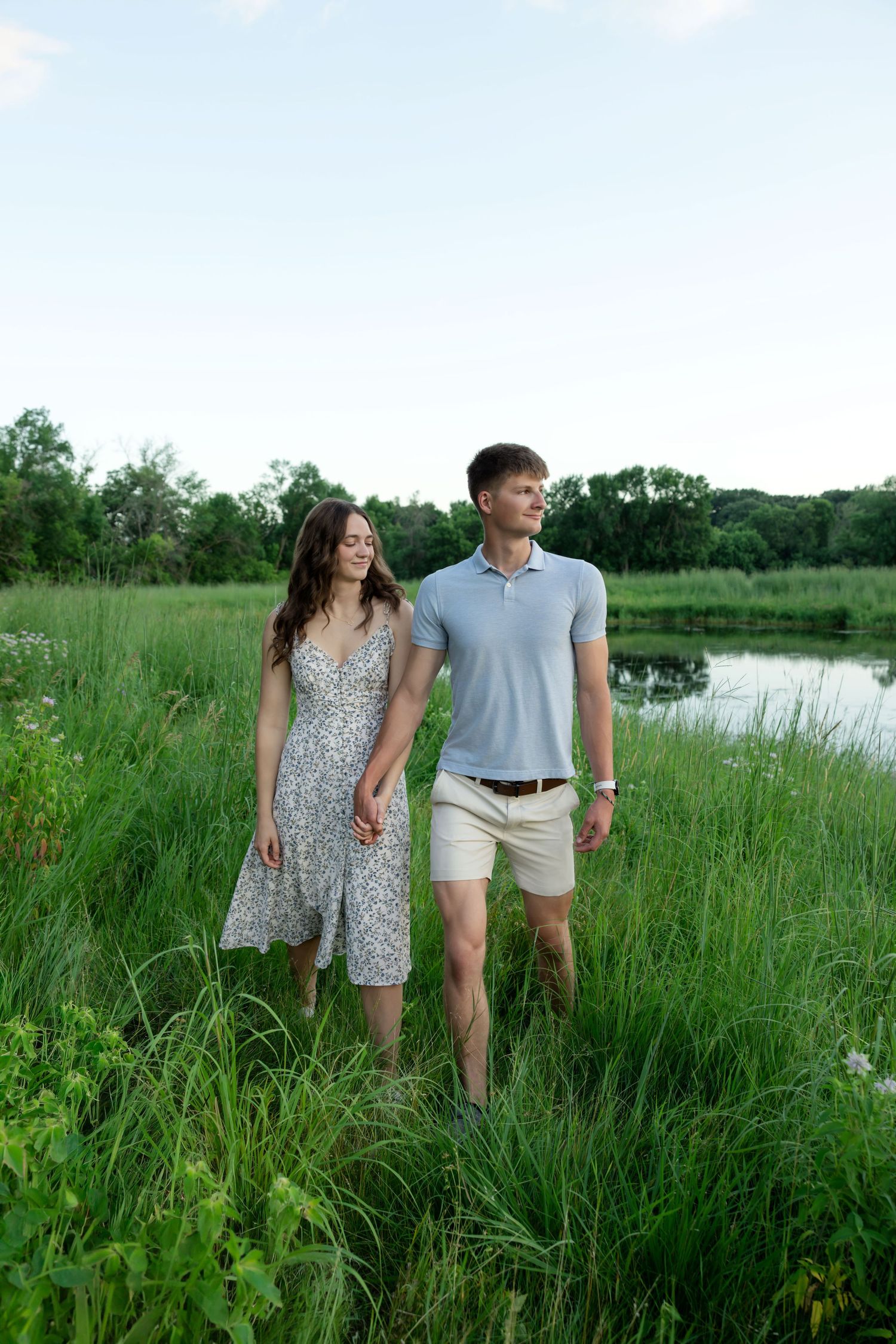 A young couple holds hands while walking through lush green grass near a pond on a warm summer evening.