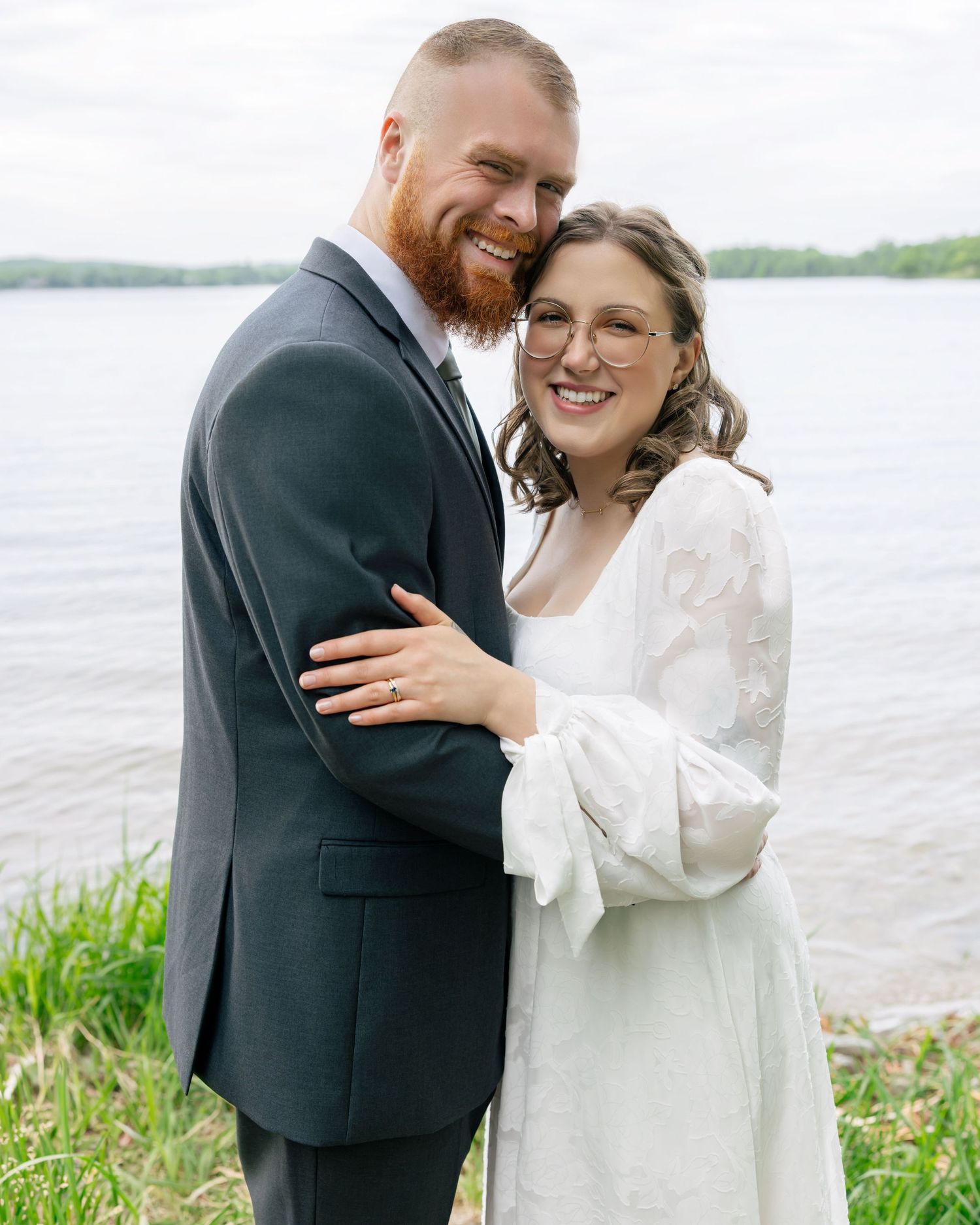 A smiling couple in wedding attire embraces near a lakeside, the groom in a dark suit and bride in a white dress with glasses.