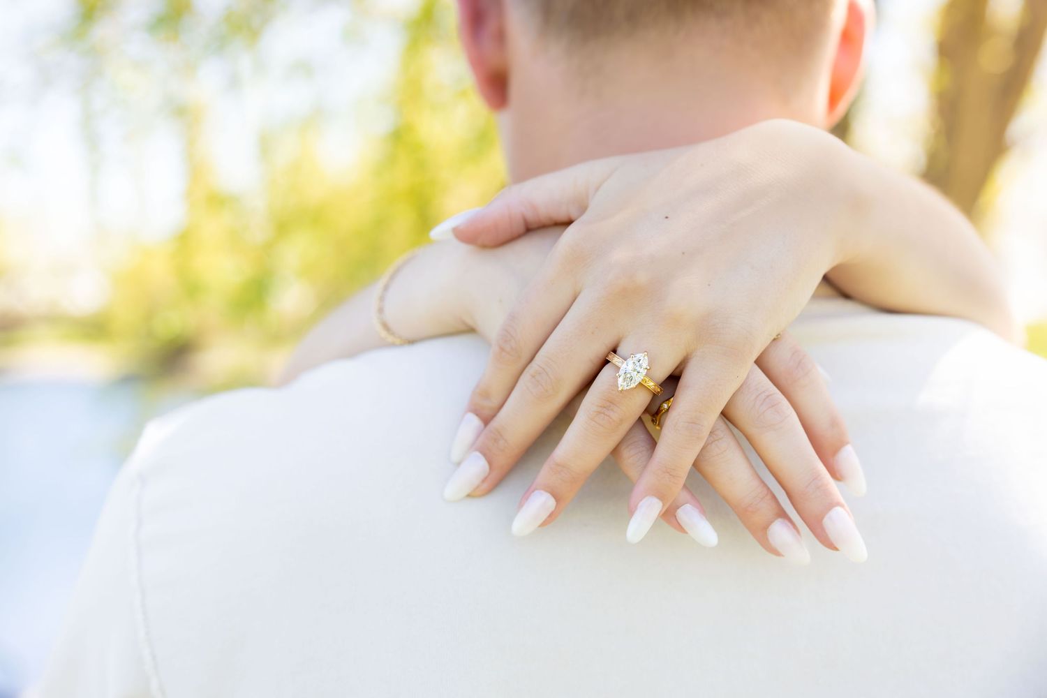 Close-up of a woman's hand with white nails and a gold diamond engagement ring resting on a man's shoulder.