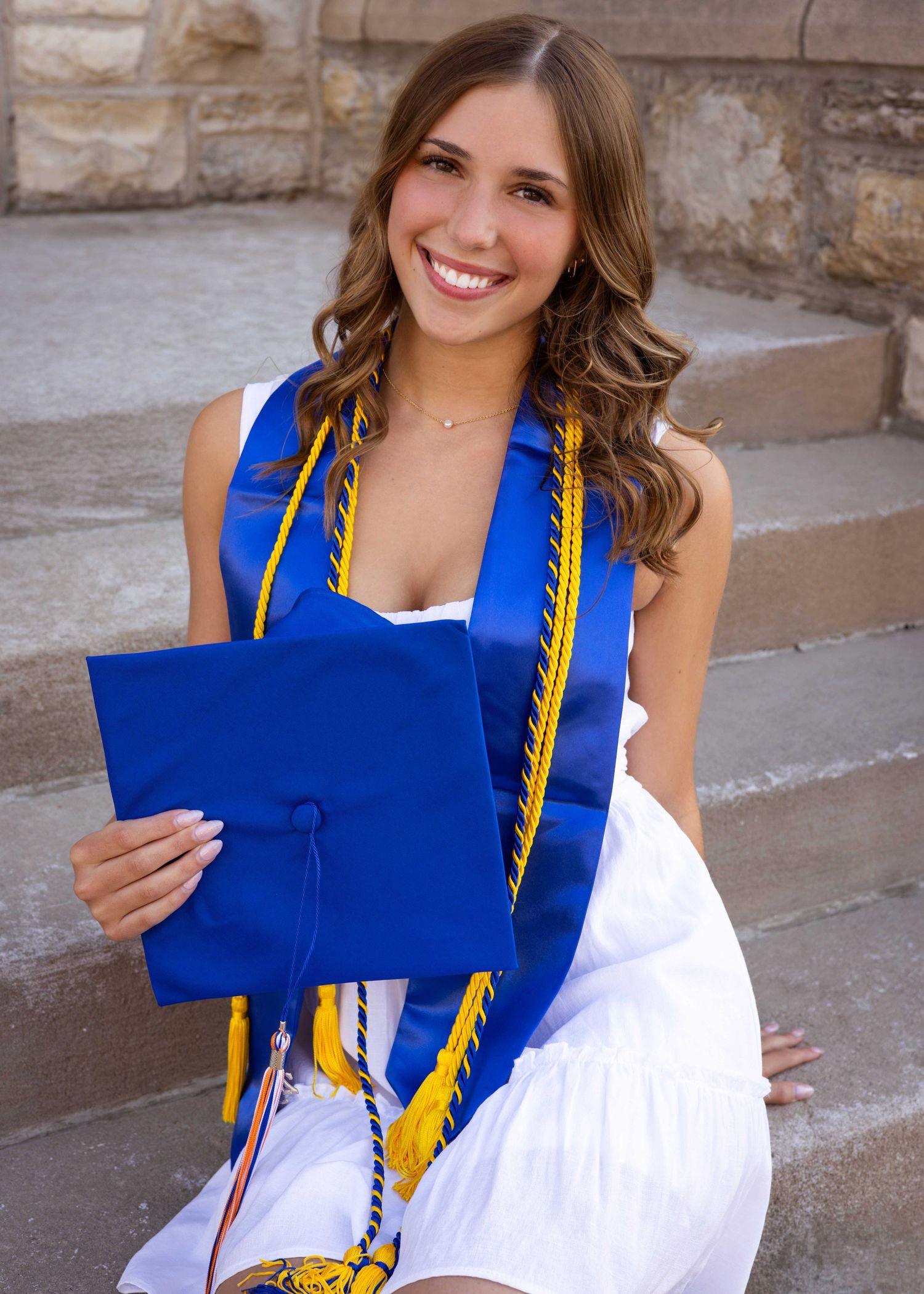 Graduate wearing honors cords holds blue diploma folder in outdoor setting.