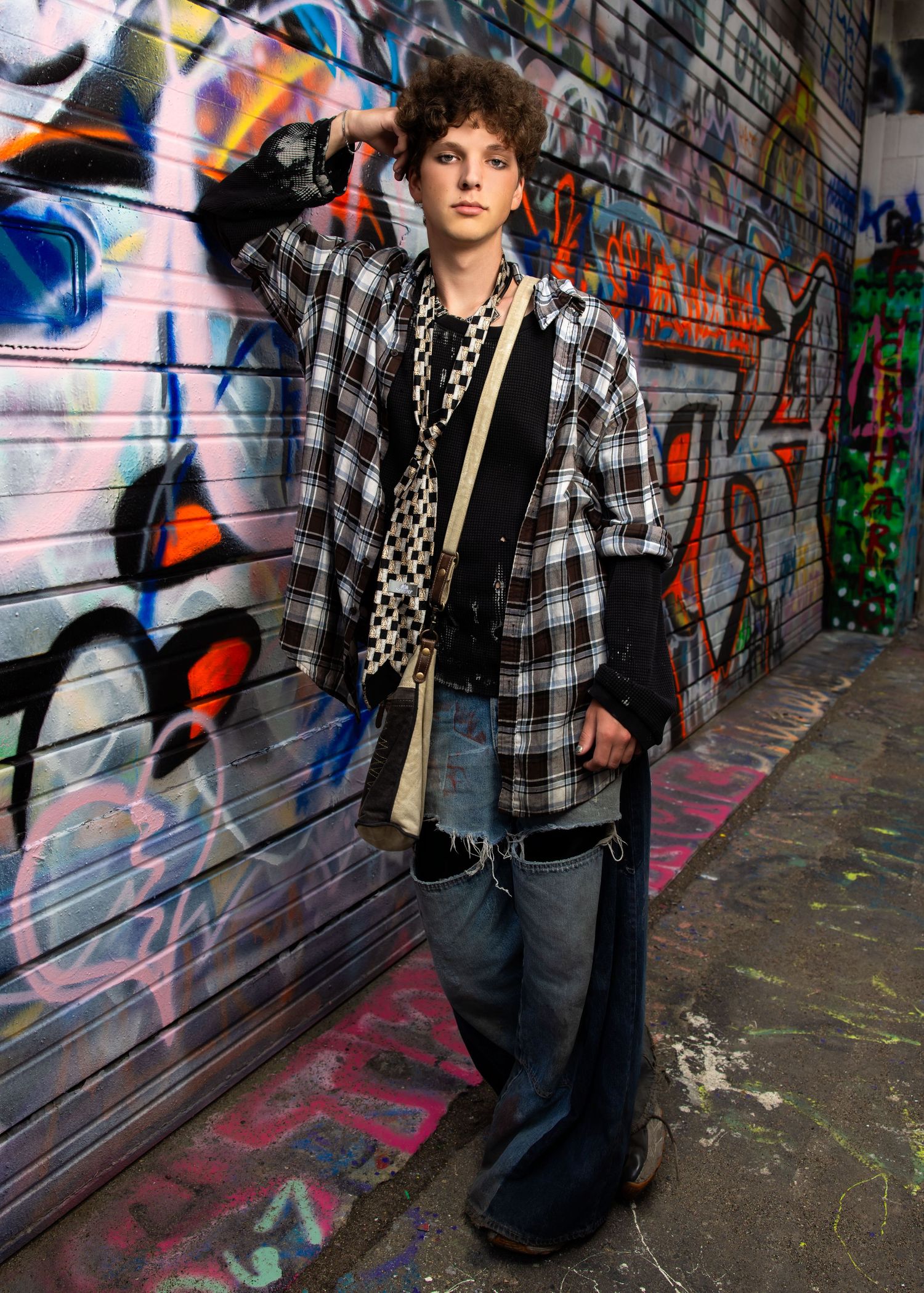 Young man in plaid shirt and wide-leg jeans posing against colorful graffiti wall in urban alley.