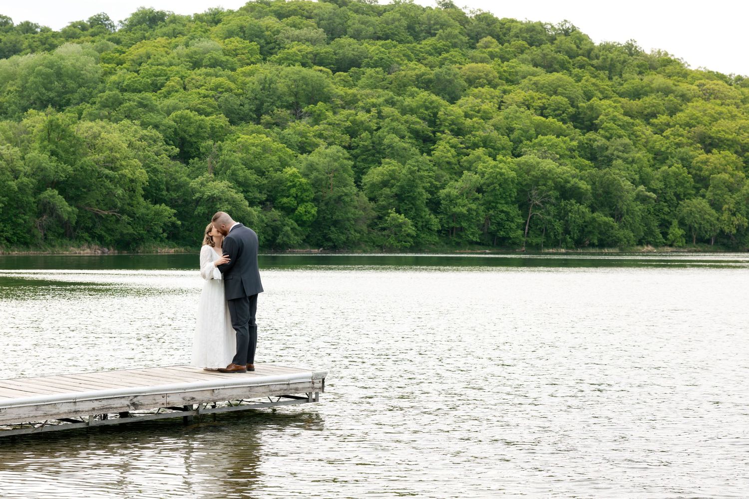 Bride and groom share a romantic kiss on a wooden dock overlooking a serene lake with lush green hills.